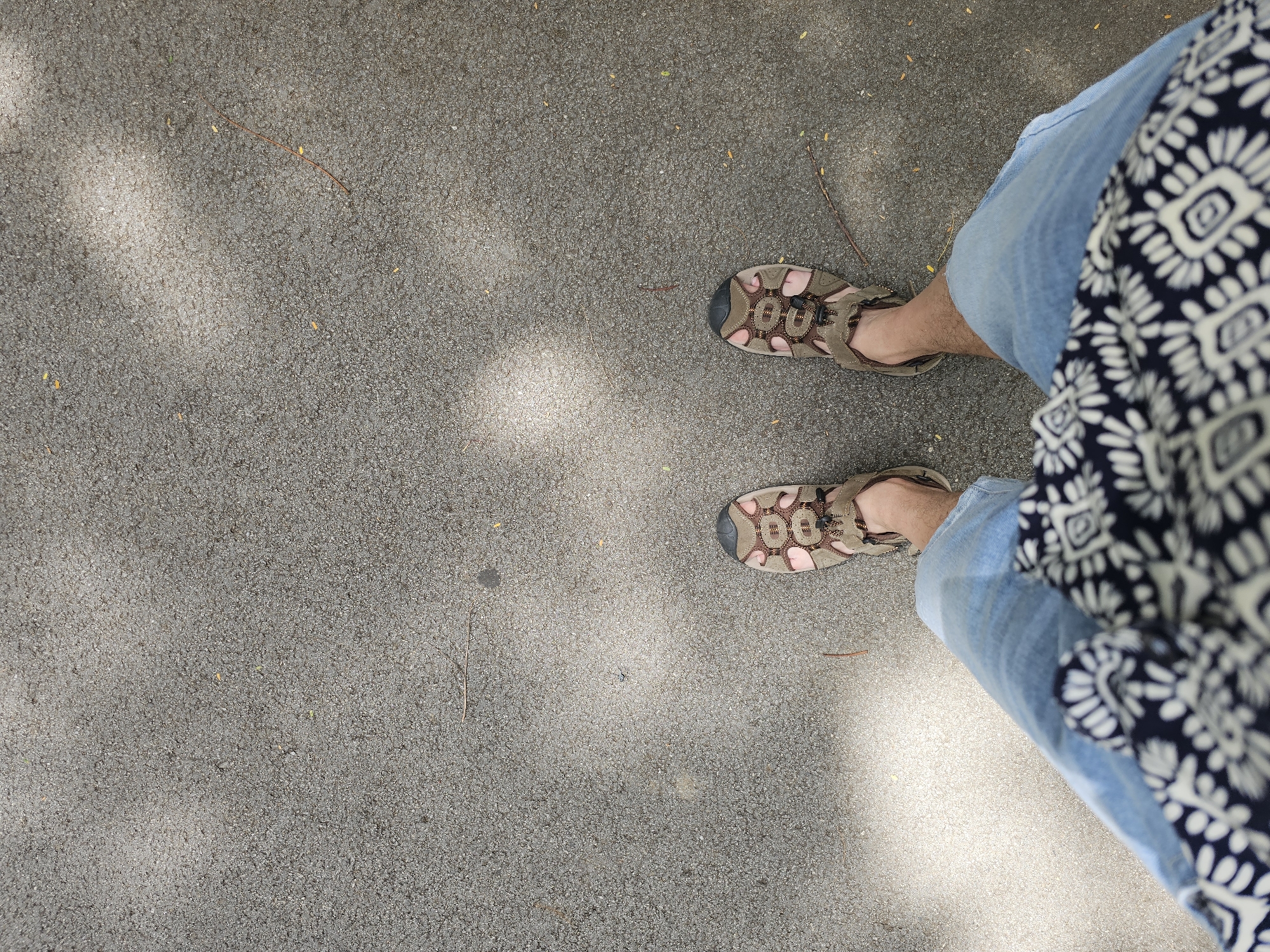 The image shows a top-down view of a person's legs and feet standing on an asphalt surface. The person is wearing light blue jeans and brown outdoor sandals. A dark-colored garment with a white patterned print is visible around the waist. The asphalt surface appears worn and has some small leaves and twigs scattered on it. Sunlight casts dappled shadows across the ground, suggesting an outdoor setting, possibly under trees. The location is not specified within the image or its context.