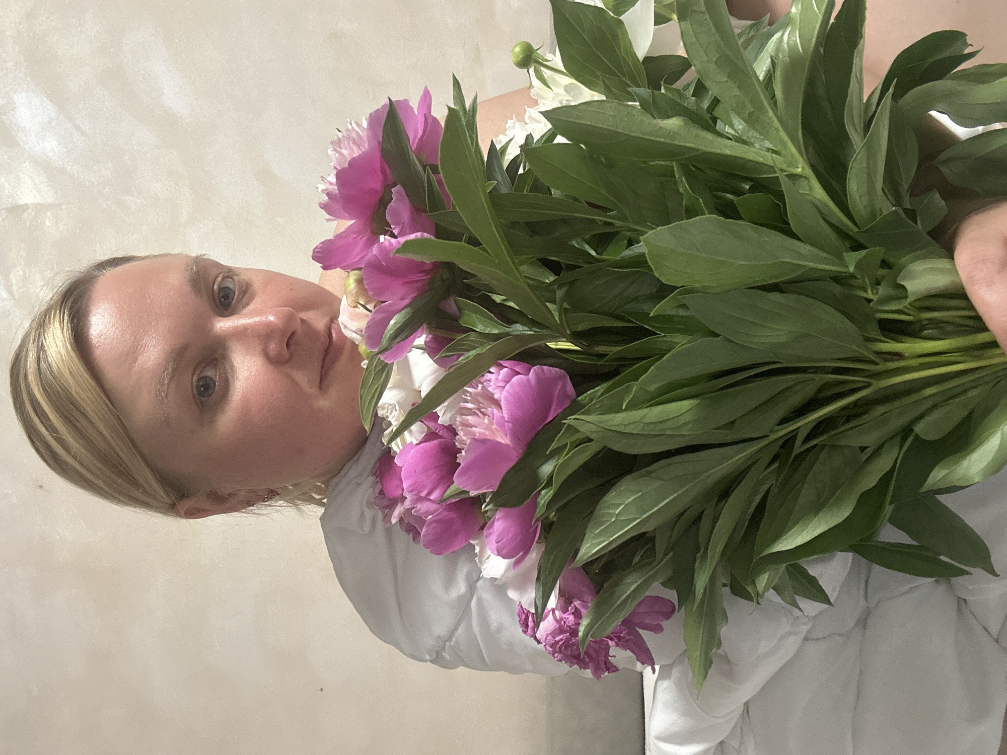 A fair-skinned woman with blonde hair tied back holds a bouquet of pink and white peonies. She is positioned in the foreground, looking towards the camera with her head slightly tilted. Her upper body is covered by a white, puffy garment, and she is seated against a textured, light-colored wall that appears to be in an indoor setting. The lighting is soft, casting a gentle illumination on the scene. The image does not provide any specific clues about the location within Chaikovsky, Russia.
