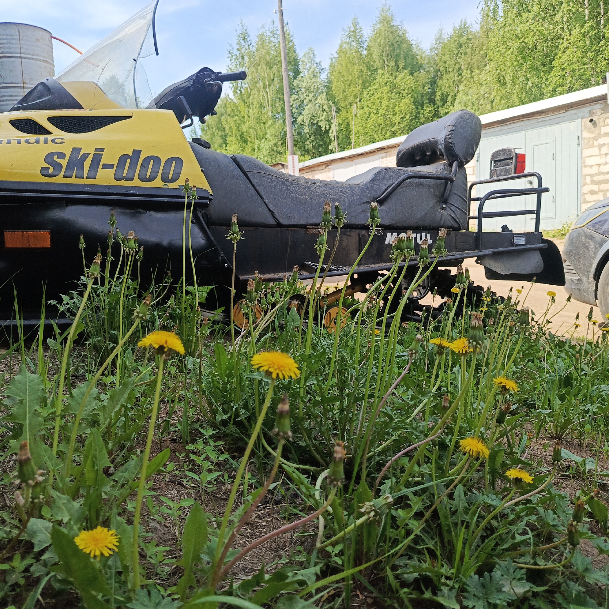 A yellow and black Ski-doo snowmobile is parked on a dirt and grass surface amidst a patch of dandelions. The snowmobile has a windshield and a black seat. Behind the snowmobile is a brick building with light green doors, and a portion of a car is visible on the right side. In the background, trees and telephone poles are visible under a partly cloudy sky. The location is in Марковский, Russia.