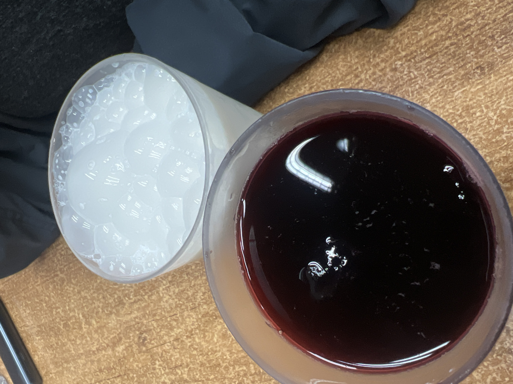 The image shows two cups on a wooden surface. One cup is filled with a white, bubbly substance, possibly soap or a similar foamy liquid. The other cup contains a dark red liquid. The environment includes a dark gray fabric item that is close to the white cup and a brown/yellow wood surface, which could be a table. The lighting is bright, causing reflections on the surface of both liquids. The exact location in Mexico City, Mexico cannot be determined from this image.