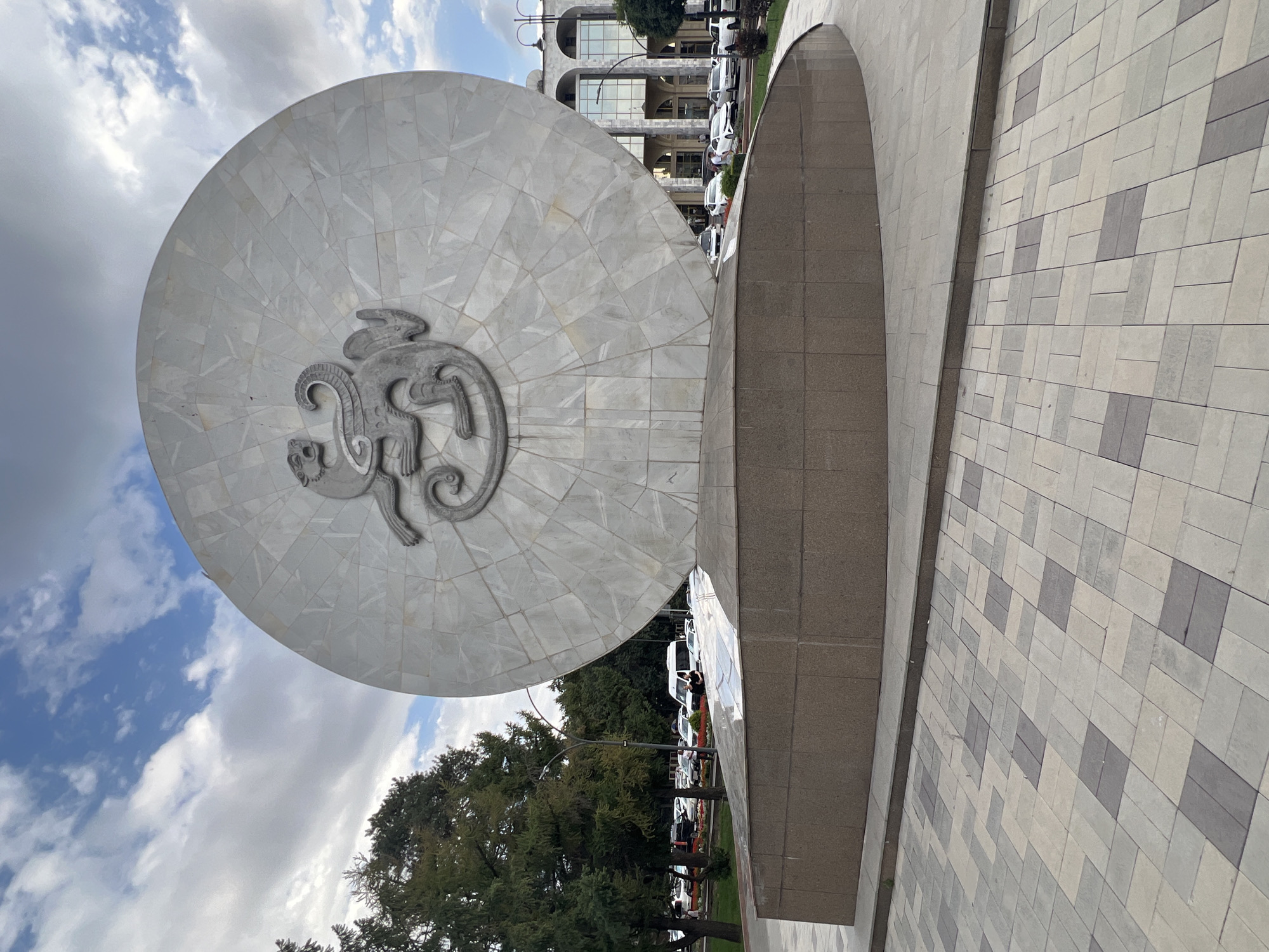 This is a full shot of the "Monument to Manas Ata" in Bishkek, Kyrgyzstan. The monument features a large, circular, light-colored stone disk with a relief of a mythical creature in the center. The disk is mounted on a tiered, brown stone base. The foreground shows a patterned paving stone walkway. In the background, there are trees, cars parked along a street, and a building with arched windows. The sky is partly cloudy.