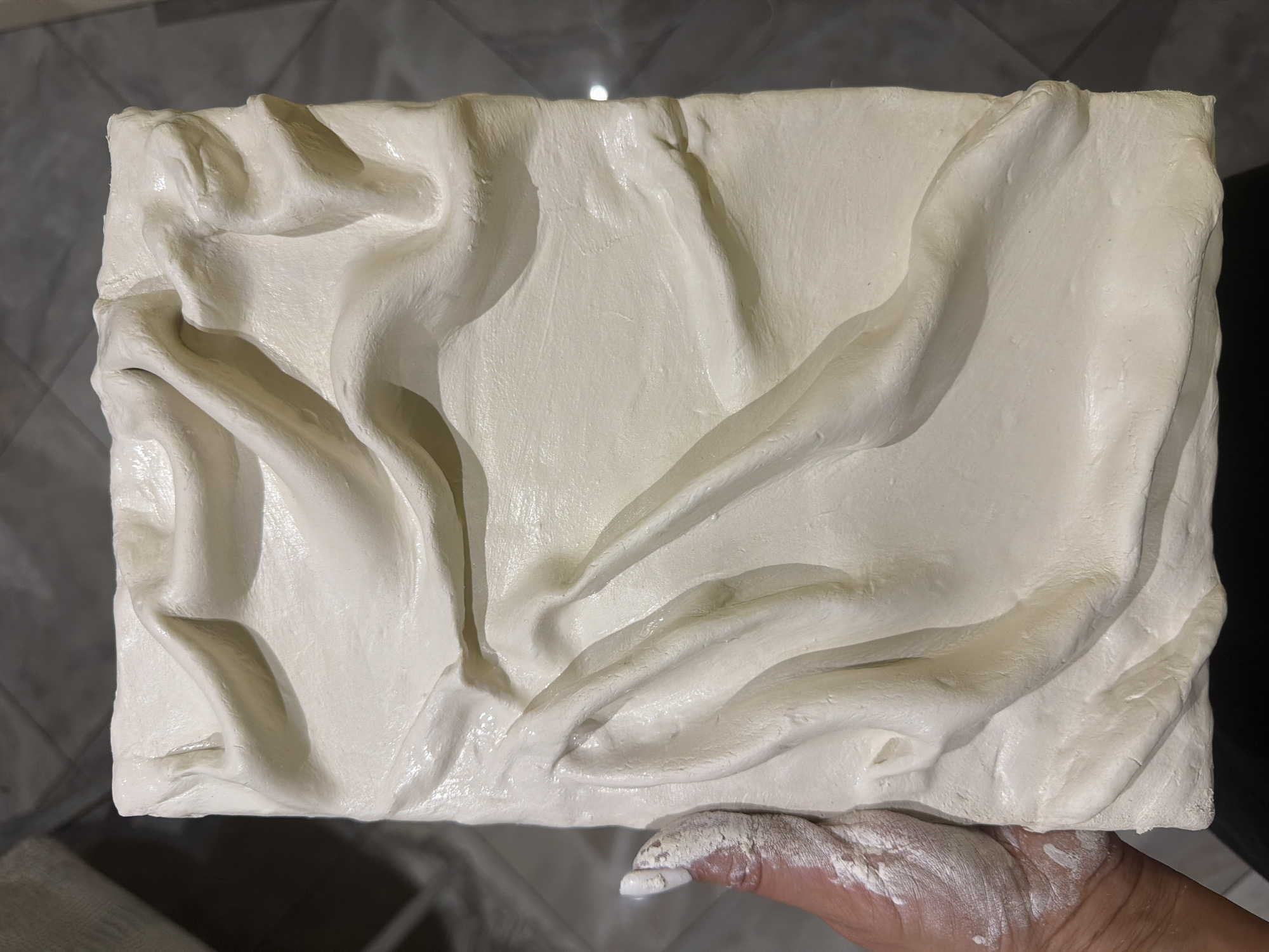 A rectangular white slab with an uneven surface of raised and recessed areas is being held up by a human hand. The slab has a shiny texture. The hand holding it is dusted with a white powder. The background is a tiled floor. The location and city are unknown.