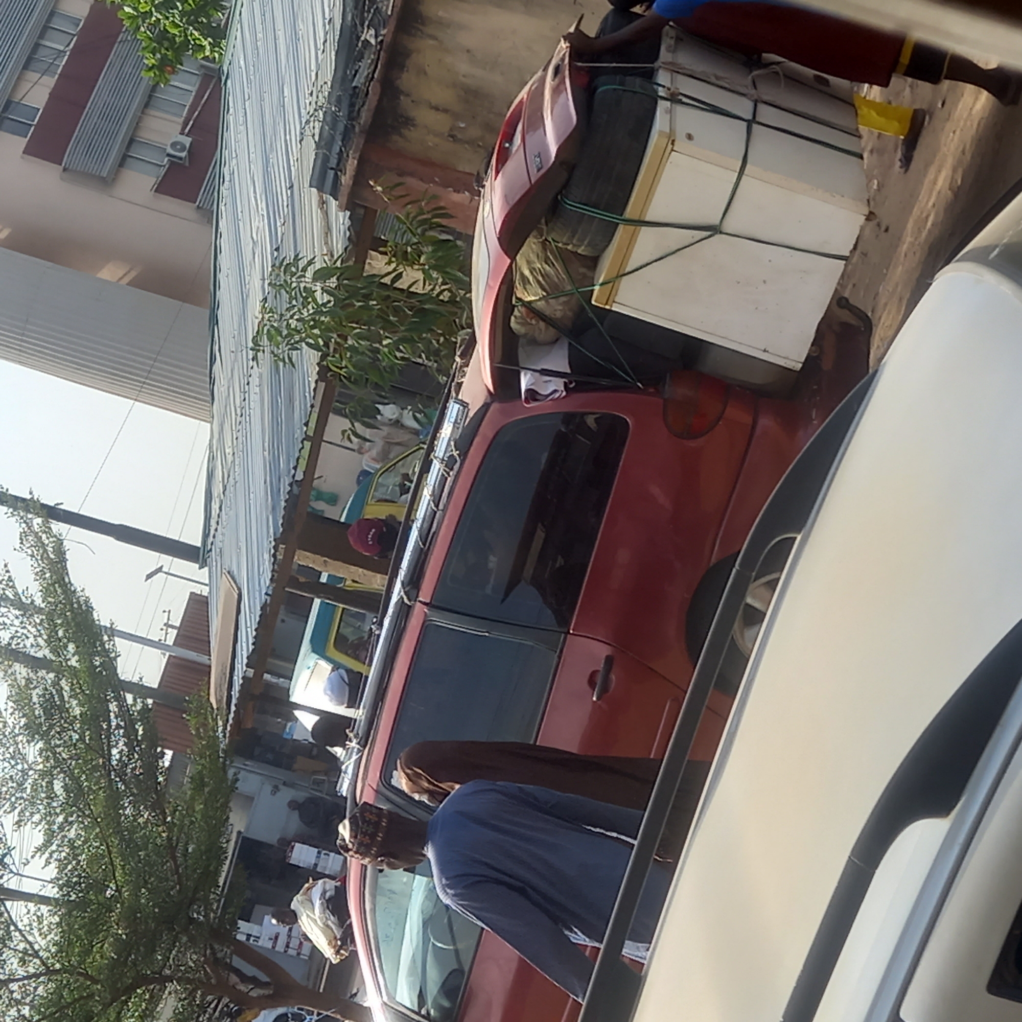 A red minivan is heavily laden with what appears to be a white refrigerator, strapped to its roof with rope, and possibly other household items. The scene appears to be an urban street setting with buildings and structures visible. Two people are near the vehicle, one is wearing a blue shirt and the other is wearing a brown hijab. The location is Dadi Riba, Nigeria, but it is not possible to determine the exact location within the city. Other cars are parked in the scene.