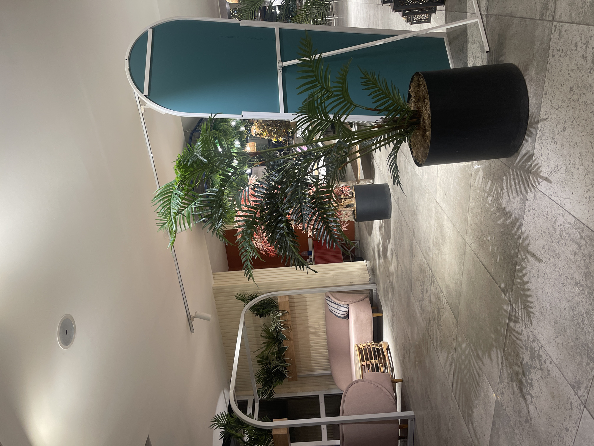 The image captures a modern, tranquil interior space, likely a commercial lounge or dining area, characterized by a contemporary design and lush artificial greenery. The setting features a light gray tiled floor and a white ceiling with recessed lighting.
In the foreground and mid-ground, several large potted plants, resembling palm trees or ferns, are prominently displayed. One particularly large plant in a matte black cylindrical pot stands on the right, casting distinct frond shadows on the tiled floor. Another smaller plant in a gray pot is visible behind it.
To the left, a curved, white-framed partition with a teal-colored panel forms a striking decorative element or partial divider. Further into the background, a cozy seating area is arranged. This area includes a light pink or beige sofa with a striped cushion and a small, possibly wicker, accent chair. This seating ensemble is partially enclosed by another arched, white-framed structure adorned with more cascading green plants. Additional foliage, some with reddish hues, can be seen deeper within the space, suggesting a tropical or botanical theme.
No people are present, indicating a quiet or empty moment in the establishment. The lighting appears artificial, suggesting either evening hours or an interior space designed to be lit predominantly by internal sources. There are no identifiable location cues within the image itself beyond the general modern interior style. No text is visible.