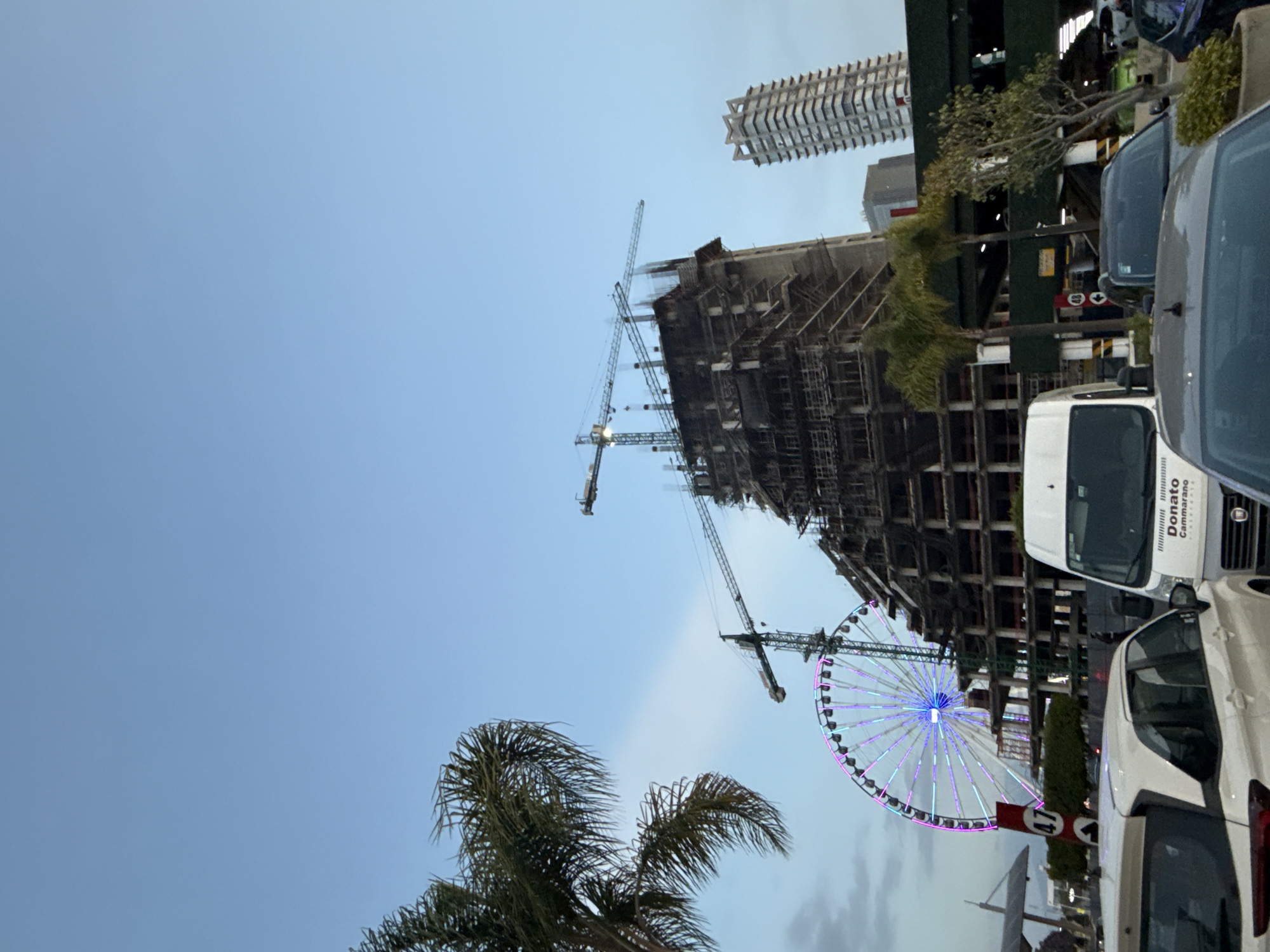 The image captures a view of Heroica Puebla de Zaragoza, Mexico, focusing on a Ferris wheel illuminated with blue and purple lights. The Ferris wheel is situated near a construction site with a partially built high-rise building, several construction cranes are visible. A palm tree is in the lower left corner, and a building with a stacked design is in the background. In the foreground, cars and a white van, marked with "Donato Cammarano" are visible on the right side.