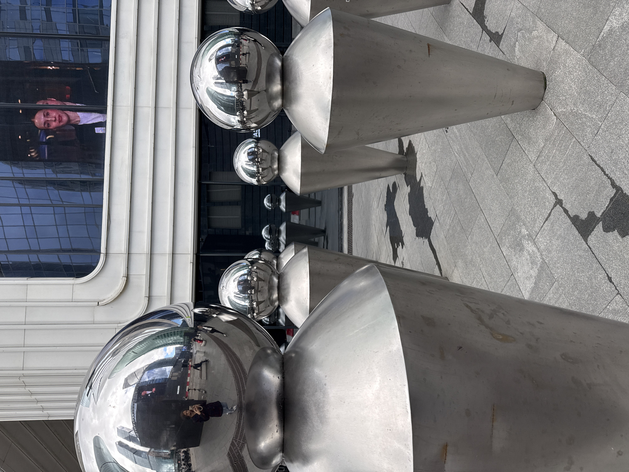 The image depicts a public outdoor space in Moscow, Russia, likely a city square or plaza, characterized by modern architecture and contemporary art installations. In the foreground, a row of large, shiny, metallic sculptures resembling stylized figures with spherical heads dominates the scene. These sculptures are arranged in a line, with the closest one partially obscuring others. The highly reflective surfaces of the sculptures mirror the surrounding environment, including a building with a large digital screen displaying two women, the sky, and other nearby structures.
The building in the background features a facade of white horizontal panels and large glass windows, with the digital screen positioned prominently. The ground is paved with grey stone tiles, some of which appear wet, creating dark patches that reflect the metallic sculptures. In the midground and background, other similar metallic sculptures can be seen, receding into the distance. A few people are visible, including a woman taking a selfie in the reflection of the nearest sculpture, and other individuals walking or standing in the vicinity. The overall atmosphere is urban and modern, with a focus on public art and architecture.