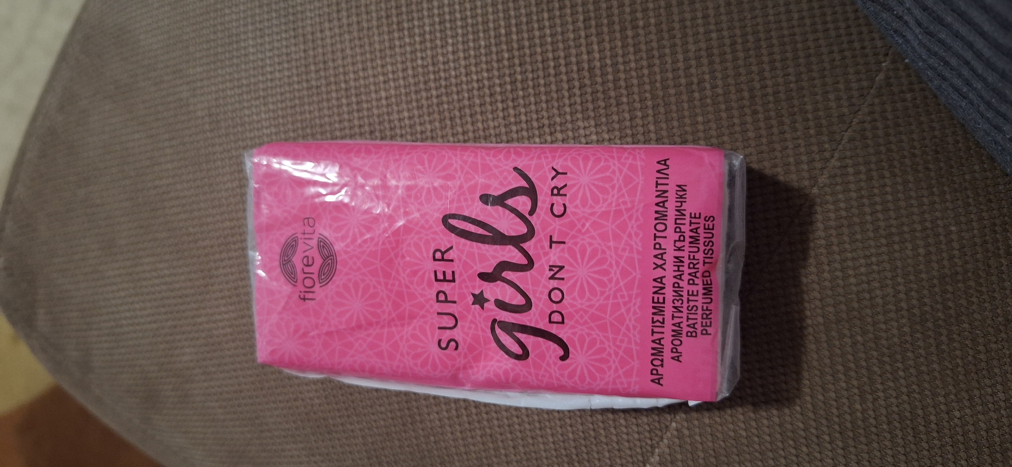 A slightly oblique, medium shot captures a sealed, rectangular packet of pink tissues resting on a textured brown surface, likely fabric. The packet is predominantly pink with a repeating pattern of small, stylized flowers in white. In bold, black script, the words "SUPER girls DON'T CRY" are printed. To the right of this text, in smaller font sizes, are lines of text in a foreign language and English, identifying the product as perfumed tissues. The brand name "fiorevita" is printed on the left in a smaller font. Some creasing of the clear plastic wrapping over the packet is visible. The scene appears to be indoors, based on the lighting and surroundings. There is no evidence that this photo was taken in Blagoevgrad, Bulgaria.