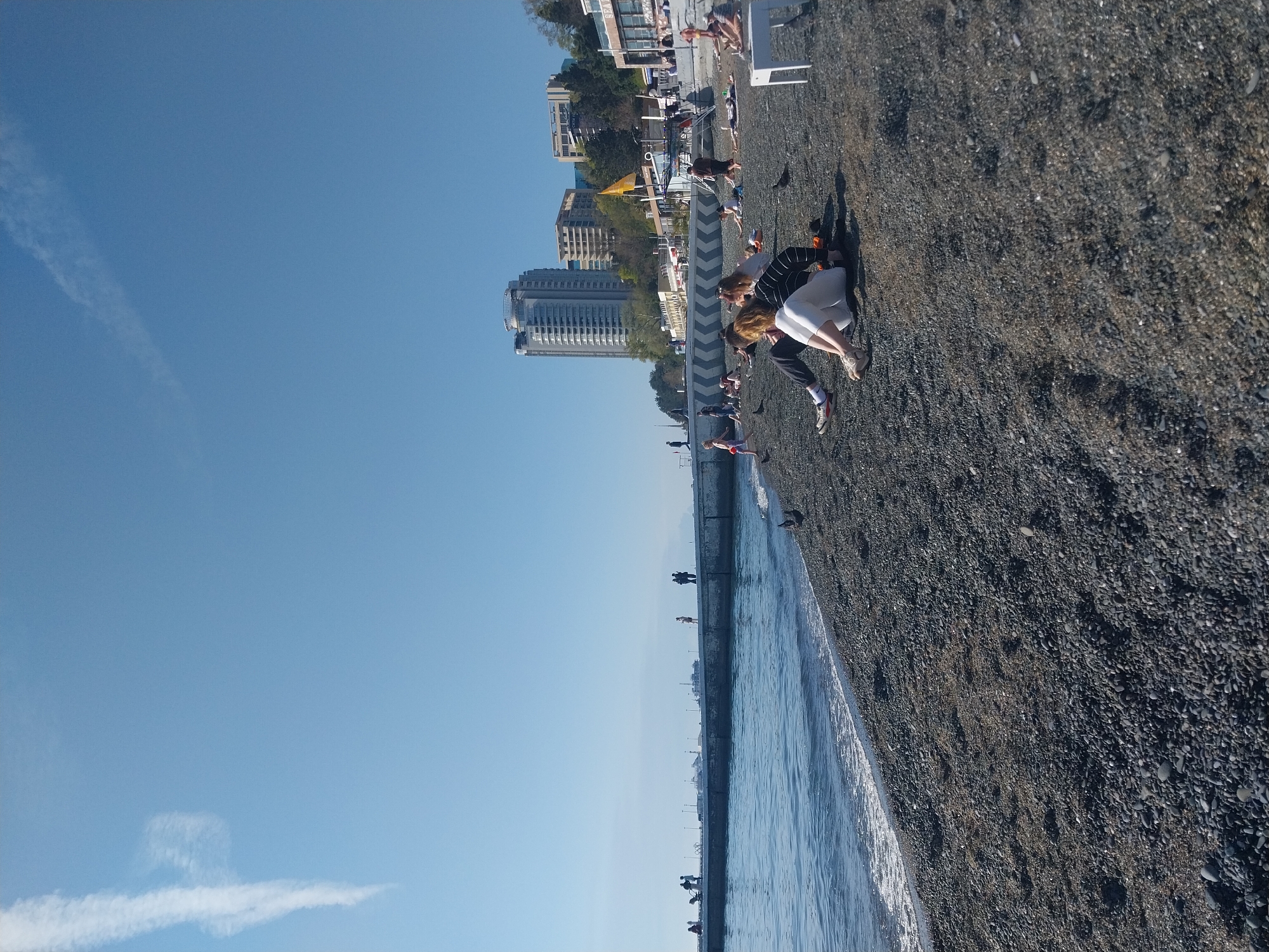 The image, oriented horizontally, presents a vibrant daytime scene at a pebbled beach. On the right, a wide expanse of dark grey pebbles stretches towards the horizon, where it meets a calm blue sea. A concrete promenade or breakwater separates the sea from the land, with gentle waves lapping against its edge.
In the foreground, towards the center-right, two individuals are crouched or kneeling on the pebbled beach. One wears dark pants and a striped top, while the other is in a white outfit, both wearing sneakers. Their attention appears focused downwards, possibly at the ground or an animal. Further back on the beach, other people are scattered, some standing, others relaxing on white sun loungers. A few birds are visible on the pebbles near the water.
Along the promenade, several distant figures can be seen walking. The background behind the beach features an urbanized coastline, showcasing a mix of modern multi-story buildings, including a prominent tall structure, and older architecture. Green trees are interspersed among the buildings, suggesting a well-established coastal town or city.
The sky above is a clear, bright blue with a faint, wispy cloud or contrail visible in the upper left. The bright sunshine indicates mid-day, casting clear shadows. No clearly legible text is discernible on any of the buildings or objects. The overall impression is one of a sunny and moderately busy seaside resort.