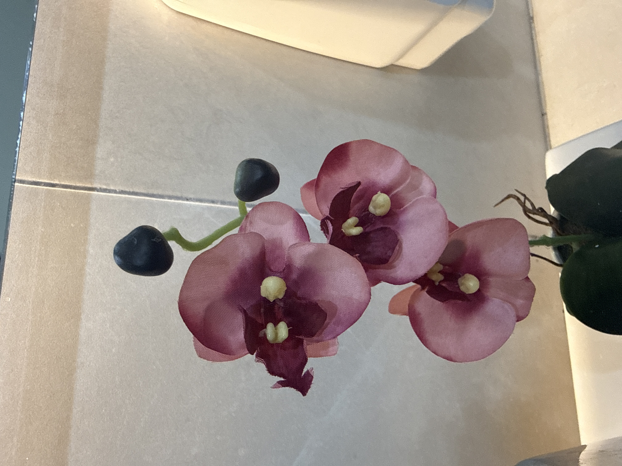 The image presents a close-up view of an artificial orchid arrangement. Three pink-hued orchid blossoms with dark purple centers and artificial yellow stamens are displayed. The arrangement includes two dark green, rounded buds on a green stem. The backdrop consists of light beige, square tiles, and a glimpse of what appears to be the bottom of a toilet paper holder in the upper portion of the image. The right side shows a glimpse of a white cylindrical object that suggests the base of the arrangement. Given the sterile and generic nature of the bathroom, there is no indication of this image being located in Mexico City. The scene portrays an artificial plant placed in a bathroom.