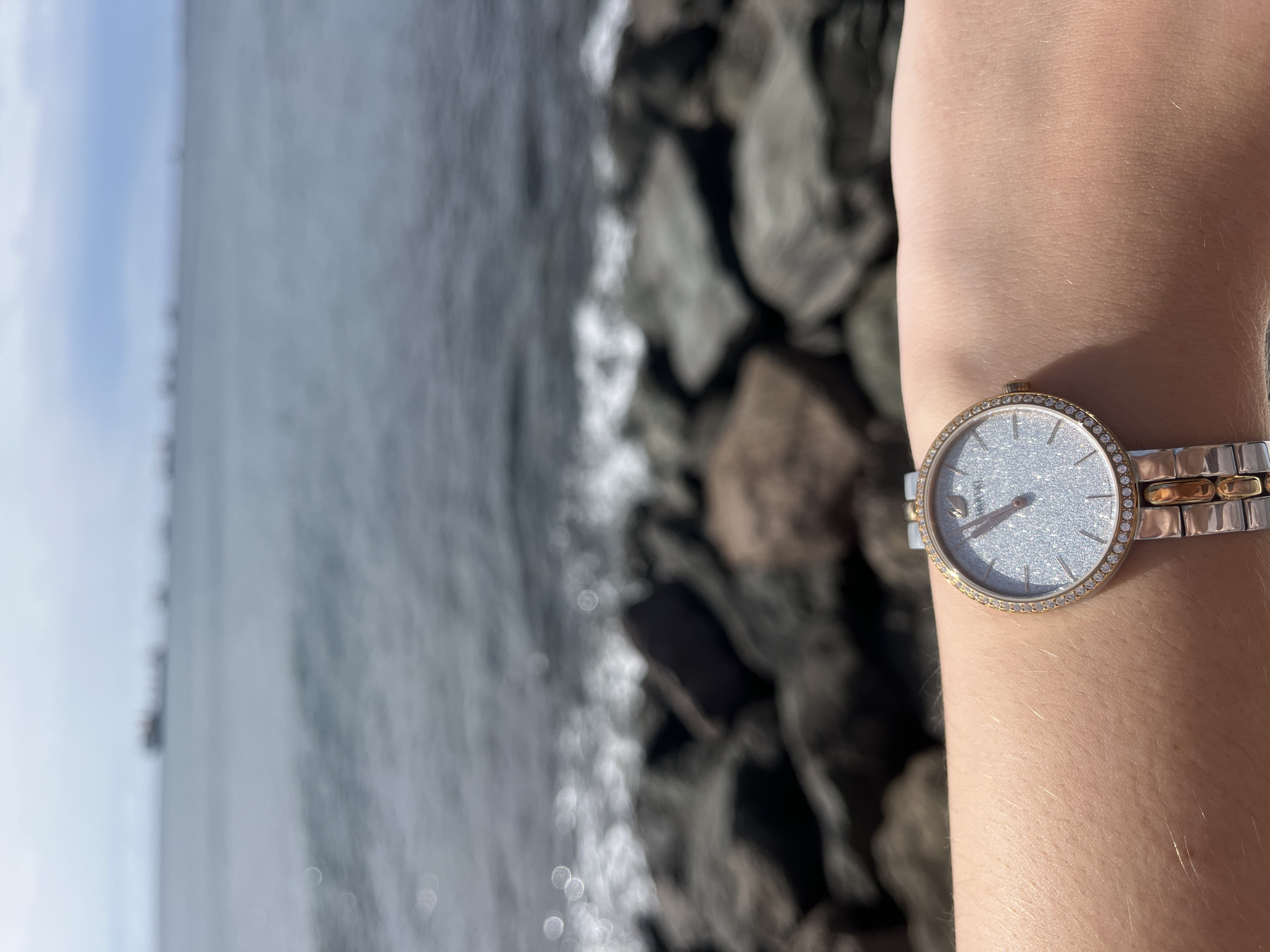 A close-up, slightly blurred photograph shows a person's arm with a silver and gold watch, against a background of the sea and rocks. The watch is on the person's left wrist, with the face showing. The sea is grey and slightly blurred, with some white wave action visible. Large, dark grey rocks form the shoreline behind the arm. The location appears to be in Las Palmas, Spain, based on the landscape. The time on the watch is approximately 10:00.