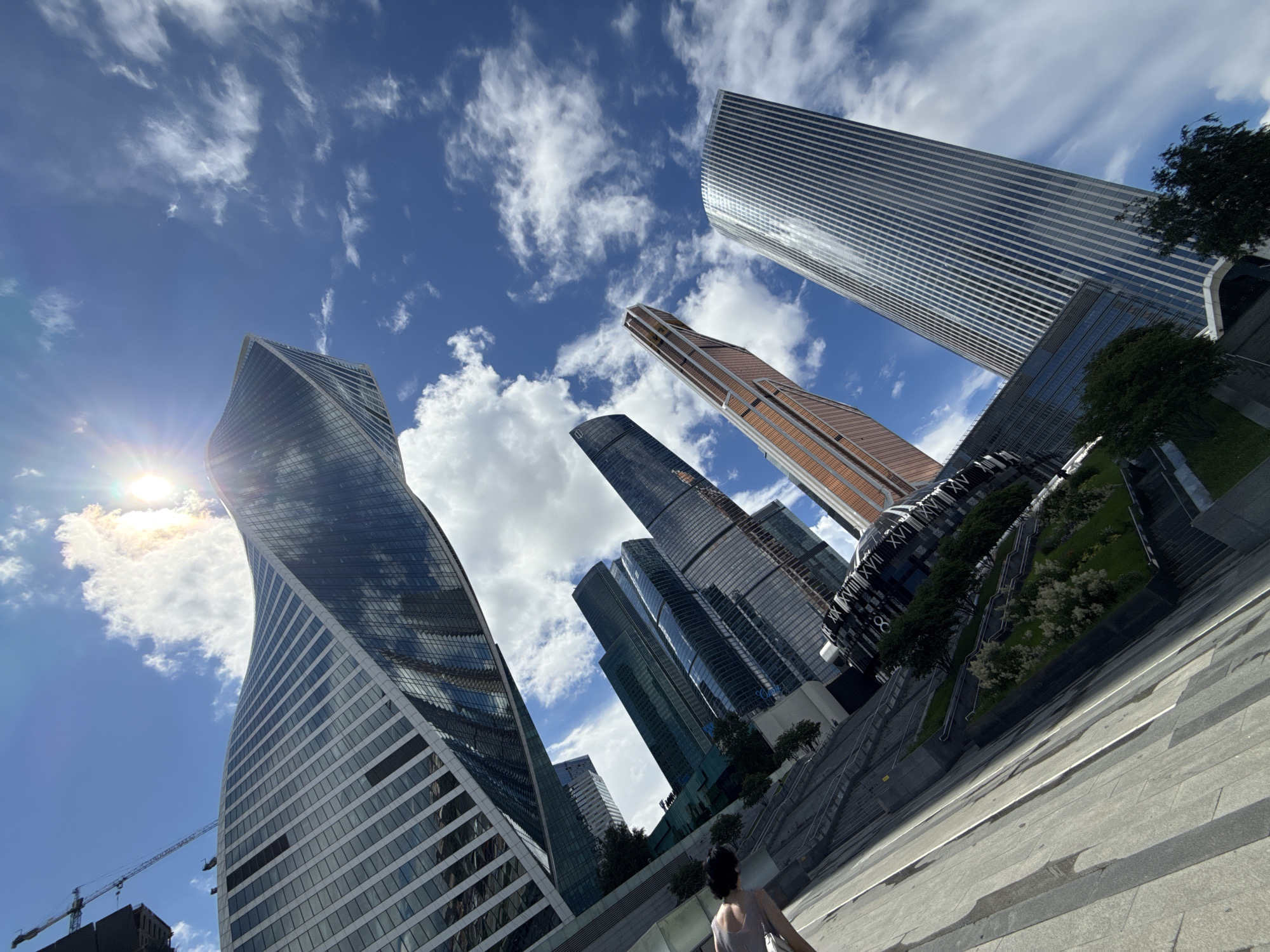 This media file is a low-angle shot of a modern urban landscape featuring several towering skyscrapers under a bright, partially cloudy sky. The sun is visible, creating a lens flare effect. The architecture is diverse, with one building featuring a distinctive twisted, glass-covered design, while others are more rectilinear with reflective facades. Green trees and landscaped areas are integrated into the urban design, suggesting a well-maintained public space. A person is visible in the foreground, walking away from the camera, adding a sense of scale to the immense structures. A construction crane is also present in the distance, indicating ongoing development in the area. The scene suggests a clear day, likely in the late morning or early afternoon, given the intensity of the sun. The overall impression is one of grand modern architecture and urban planning in a bustling city. While no specific text is legible on the buildings, the architectural style is characteristic of major metropolitan centers. The location is Moscow, Russia, and the skyscrapers likely belong to the Moscow City business district.