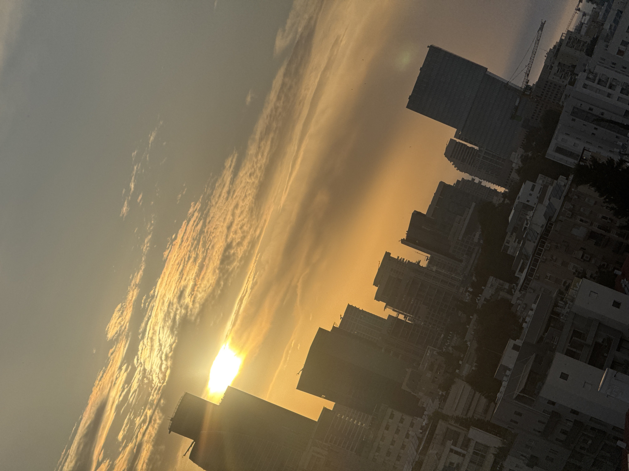 This media file captures a dynamic urban sunset in Ramat Gan, Israel, from an elevated vantage point. The scene features a sprawling cityscape composed of numerous tall buildings and modern high-rises, primarily silhouetted against a brilliant sky.
The sky is a central element, showcasing a dramatic blend of warm golden-orange, yellow, and soft grey tones. Streaky clouds are intensely illuminated by the low, bright sun, creating a strong contrast with darker cloud patches. The sun itself is a powerful, overexposed orb positioned low on the horizon, casting a strong backlight across the scene and producing a noticeable lens flare.
Among the densely packed architectural structures, a prominent construction crane is visible, indicating ongoing urban development. The buildings exhibit various contemporary designs, with some details of windows and balconies subtly discernible despite the heavy shadows. The overall impression is one of a bustling city transitioning into night, bathed in the rich glow of dusk.
No people or specific activities are explicitly visible, and there is no discernible text in the image. The photograph is composed at a dynamic angle, enhancing the perspective of the cityscape and the vastness of the sky.