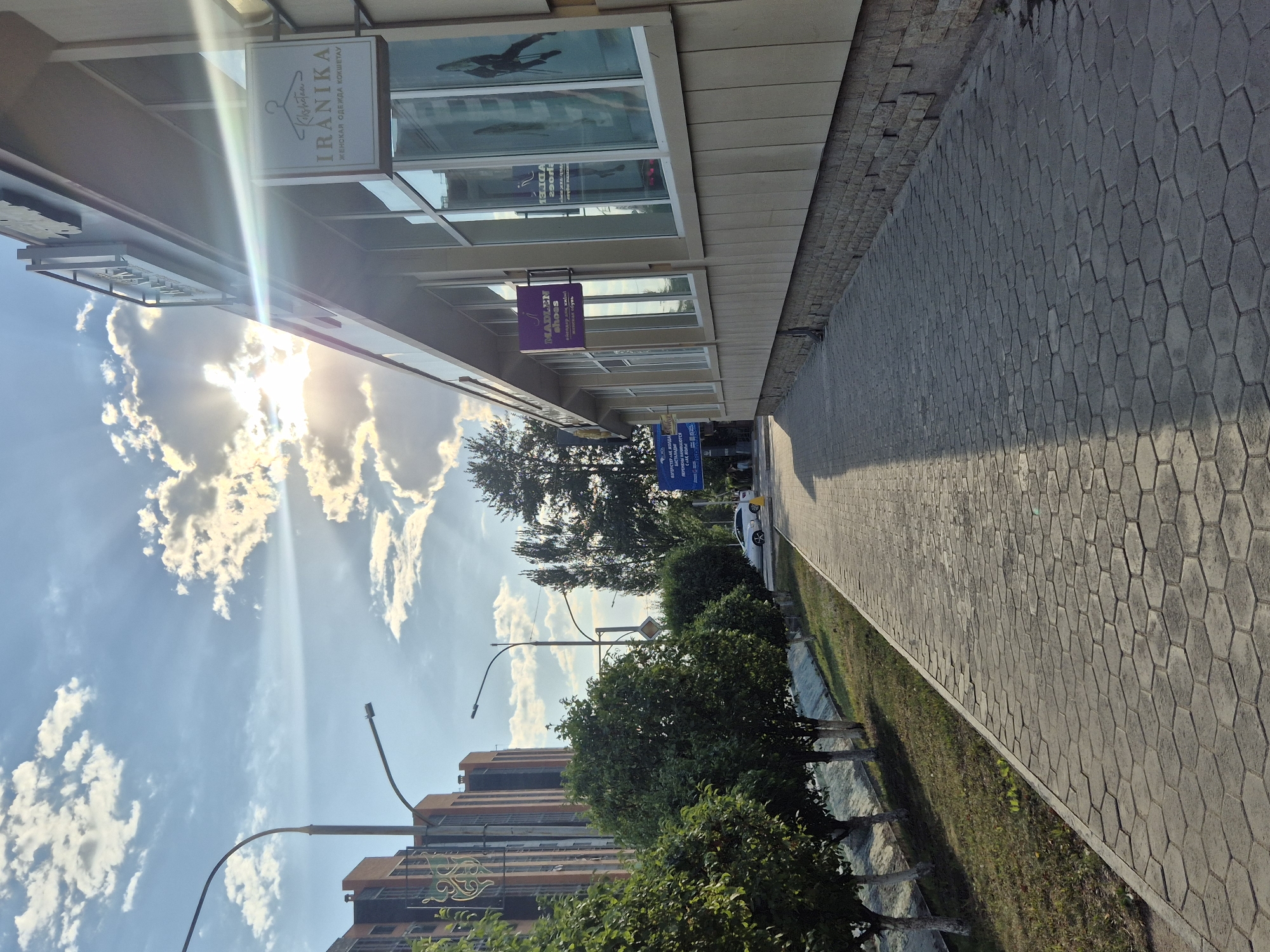 The image depicts a street view in Kokshetau, Kazakhstan, under a bright, partly cloudy sky. In the foreground, a paved pedestrian walkway, constructed with hexagonal grey stones, stretches along the right side of the frame. To the left of the walkway, a strip of green grass borders a shallow, dry canal. Several trees with white-painted trunks are planted along the grass.
Adjacent to the walkway is a low-rise building with a beige, horizontally paneled facade and a stone-clad foundation. The building has large windows. A prominent sign reading "IRANIKA" with an image of a hanger and the text "ЖЕНСКАЯ ОДЕЖДА КОКШЕТАУ" (Women's Clothing Kokshetau) is mounted on the building's exterior. Further down the building, another sign, purple and rectangular, reads "MADLEN shoes". Reflections of the sky and trees are visible in the building's windows.
In the background, on the left side of the image, a tall, modern residential building with orange and dark grey elements stands. Streetlights with curved poles are visible along the street. A white car is parked on the street. The sun is shining brightly, casting long shadows across the walkway and creating sun flares in the sky. The overall scene suggests a daytime view of a commercial and residential area in Kokshetau.