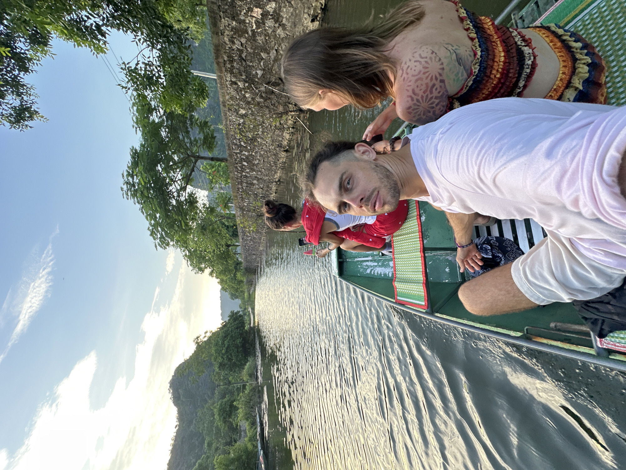 A long shot captures three individuals on a boat traveling down a river. The person in the foreground, a man, faces the camera, his gaze direct. Behind him, two women are seated. The woman on the left is wearing a red top with a white undershirt, while the woman on the right faces away from the camera, displaying a distinctive tattoo on her back. The boat, painted green, moves through calm, rippling water reflecting the sky. The surrounding environment is lush, with verdant trees and hills flanking the river. A stone wall runs along the right bank of the river, with trees growing from it. The sky above is a blend of blue and white clouds, with the soft light suggesting late afternoon or early morning. The scene appears to be set in Nam Hoa Lư Ward, Vietnam, a region known for its natural beauty and waterways.