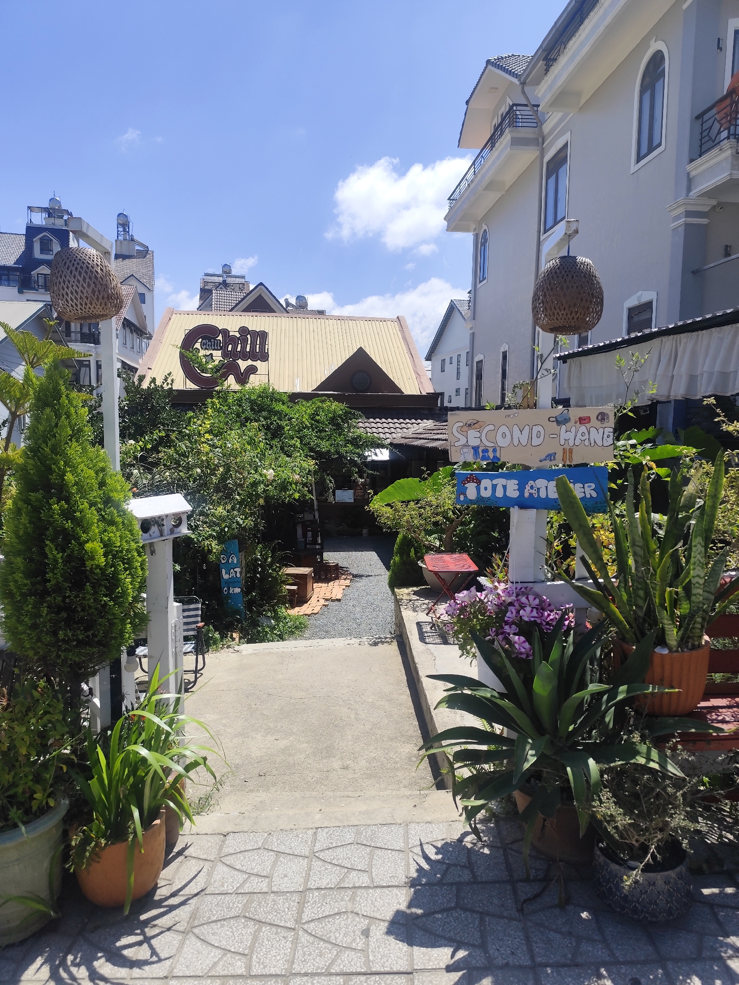 The image shows a narrow pathway leading through a lush garden towards a building with a yellow, sloping roof. A prominent sign for "Chill" is visible above the entrance. To the right, a white post displays two smaller signs: "SECOND-HAND" with illustrations of bags and shoes, and "TOTE ATELIER" with mushroom graphics. Various potted plants, including snake plants and flowering varieties, are arranged along the pathway and in front of the buildings. The architecture suggests a residential area in Da Lat, Vietnam, characterized by houses with pitched roofs and decorative elements. The sky is blue with scattered clouds, and the scene is bathed in daylight, indicating an outdoor setting during daytime.