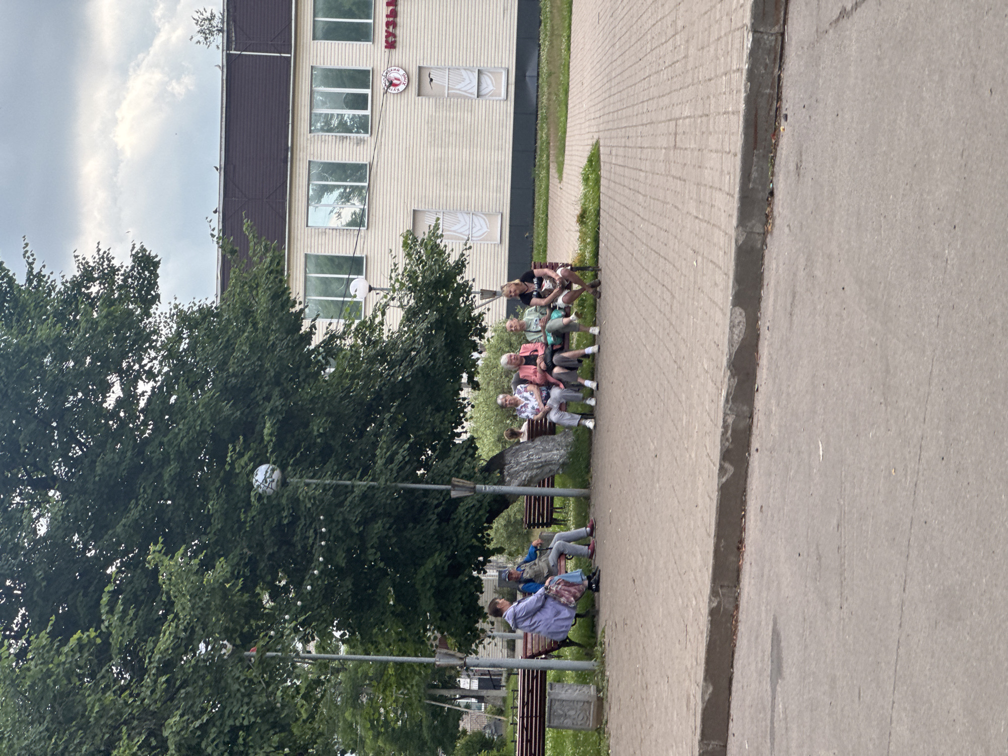 In Kuzmolovskiy, Russia, several people are sitting on park benches in front of a light-colored building. A large tree with green leaves is in the foreground, casting shade. The building has multiple windows and a sign with Cyrillic text. The ground is paved with brick-like patterns. One person is wearing a blue jacket, and others are dressed in casual attire. The sky is partly cloudy.