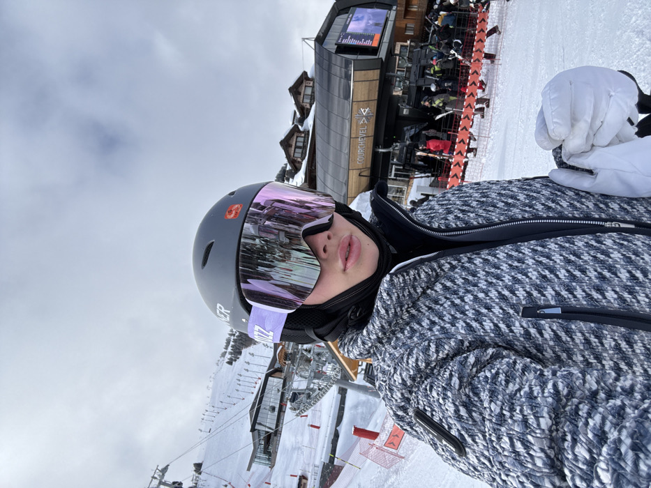 A close-up, eye-level photograph captures a person in ski gear posing for a selfie or portrait on a snowy slope. The individual wears a gray ski helmet with "NER" visible and a red circular sticker, oversized mirrored ski goggles with purple and pink tints reflecting the environment, a black balaclava, and a textured black and white ski jacket, possibly a herringbone pattern, with visible zippers. A white ski glove is held in the lower right foreground.
The background reveals a bustling ski resort in Courchevel, France. Directly behind the subject to the right is a modern ski lift station with "COURCHEVEL" prominently displayed, along with a symbol. A queue of skiers and snowboarders in various colored apparel is visible, waiting to board the lift. To the left, a wide snow-covered ski slope ascends, marked with multiple ski lift pylons and cables. A large, contemporary building with expansive windows is situated mid-slope. The sky is overcast, indicating a cloudy day. Orange safety fencing with white directional arrows is present on the snow in the midground.