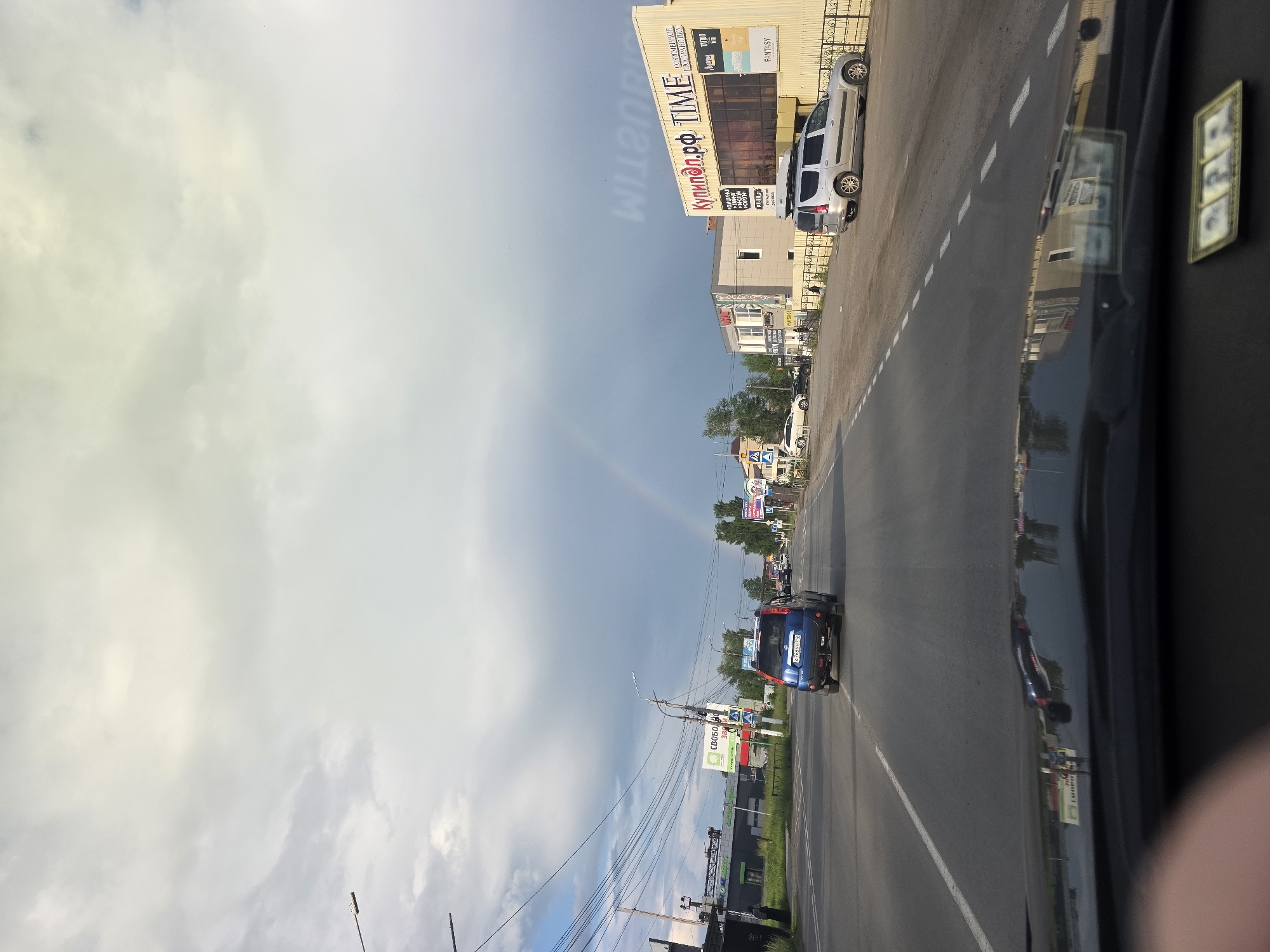 The image depicts a view from a vehicle driving on a road in Chaikovsky, Russia, under a cloudy sky with a faint rainbow visible. The road is asphalt with clear lane markings. On the left side of the road, there are buildings with advertisements and power lines. A blue SUV is in front of the vehicle, and other cars are further down the road. On the right side of the road, there are several buildings, including one with the sign "КупиЛоп.рф TIME" and another smaller building next to it. A silver minivan is parked on the right side of the road. Trees are visible in the background. The dashboard of the vehicle is in the foreground, with a small decorative item containing three photos.