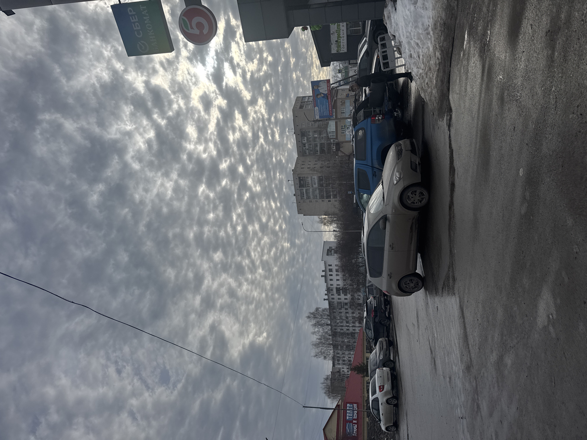 The image shows a street scene in Chaikovsky, Russia under a cloudy sky. Cars are parked along the right side of the wet street. Buildings of varying heights line the background. On the left are signs for Sberbank and a "Pyaterochka" supermarket. A person is standing near a blue truck, appearing to be interacting with the vehicle. Snow is visible along the edges of the road.