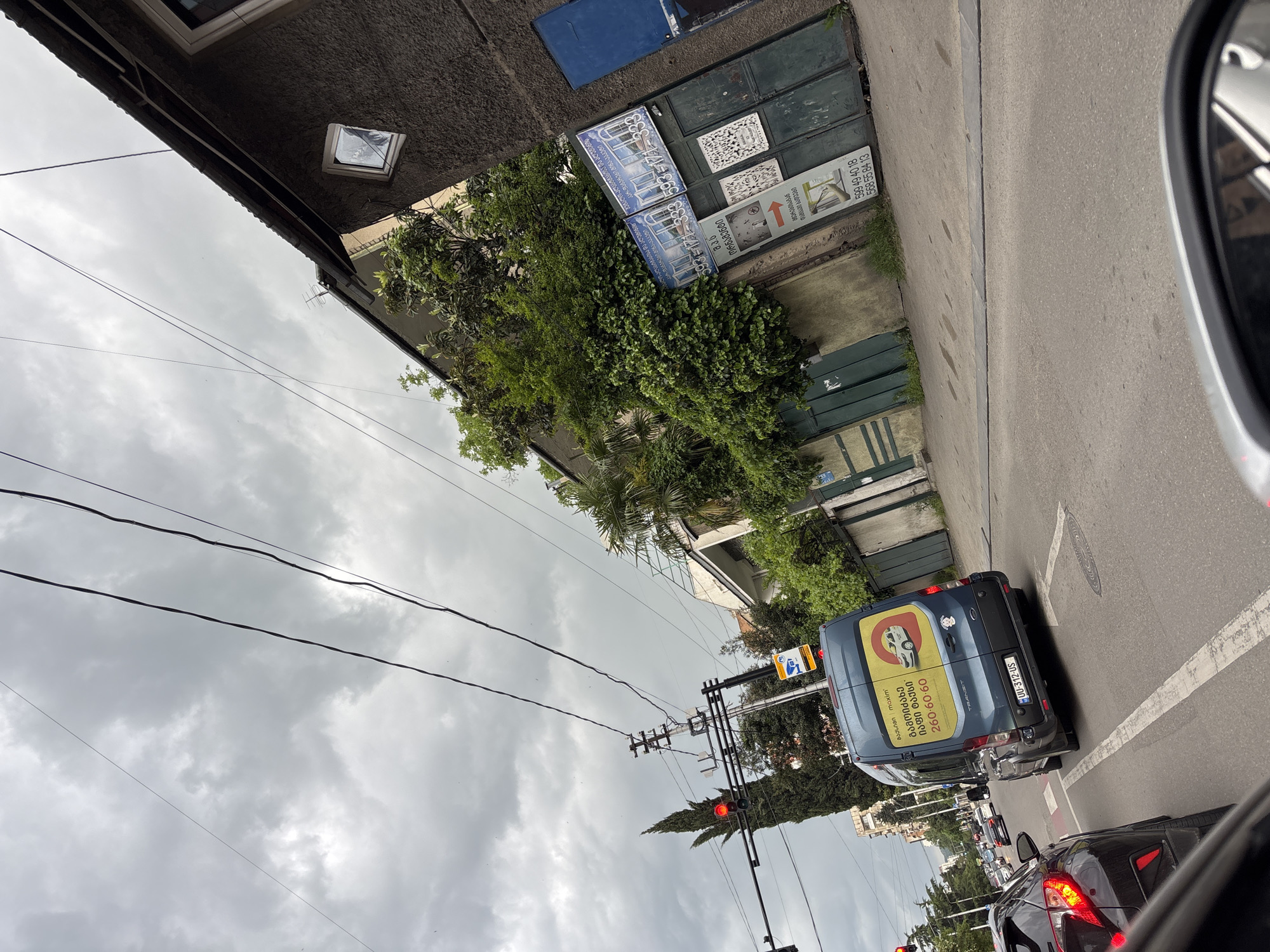 The image shows a street scene in Tbilisi, Georgia, taken from a vehicle. A blue van with a yellow advertisement for a taxi service (260-60-60) is visible in front. The license plate of the van is UU-312-US. In the background, there is a building with advertisements for metal-plastic and aluminum window and door services, including phone numbers (593-171-333, 599 49 40 18, and 568 55 84 13). The street is paved and has other cars stopped at a traffic light. Power lines are visible against an overcast sky.