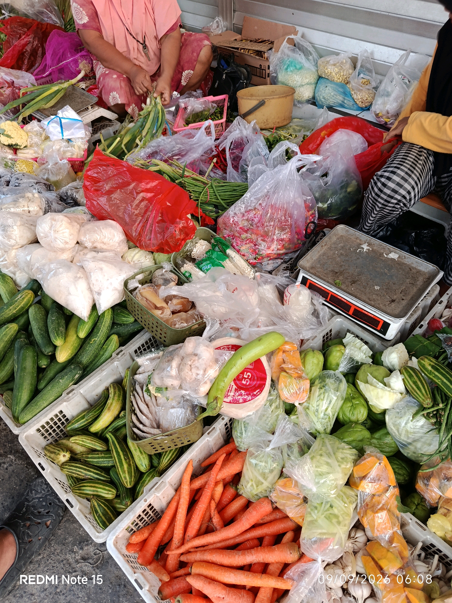This is a close-up, eye-level outdoor shot of a market stall in Keboansikep, Indonesia, with a focus on a variety of fresh produce and packaged goods. Two individuals are partially visible in the frame. On the left, a person wearing a pink and white floral patterned shirt and skirt is seated, actively sorting green onions. Their hands are engaged in the task, with a key hanging from a necklace. To the right, another person, wearing a yellow shirt and checkered pants, is also seated and appears to be interacting with the produce.
The stall is densely packed with items displayed in various containers, including white plastic crates and woven baskets. Dominating the foreground and midground are an abundance of fresh vegetables such as cucumbers, long beans, carrots, and what appear to be gourds or chayote. These are interspersed with packaged goods, including white powdery substances in plastic bags, red plastic bags containing produce, and clear plastic bags filled with various vegetables and potentially dried goods. A vintage scale with a dusty metallic surface and an orange digital display is prominently positioned on the right side of the midground. The ground beneath the stall is a rough, dark paved surface. The background is blurred, showing more market stalls and structures, suggesting a bustling marketplace environment. The image is dated "09/09/2026 06:08" in the lower right corner and is watermarked "REDMI Note 15" in the lower left.
