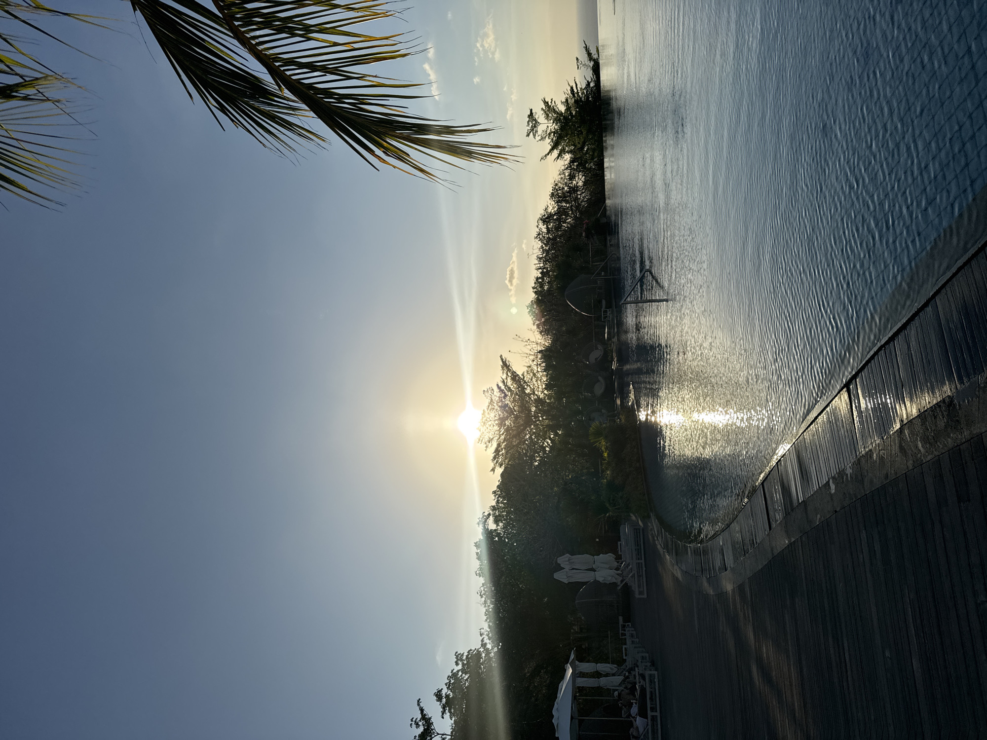 The image depicts an infinity pool at sunset in Kuta Selatan, Indonesia. The sun is low on the horizon, casting a warm glow across the sky and reflecting on the water. Palm fronds hang from the right side of the frame, framing the scene. In the foreground, a wooden deck curves around the pool, with lounge chairs and umbrellas set up on one side. Lush vegetation borders the pool, and in the distance, the ocean is visible. The overall atmosphere is serene and tranquil.