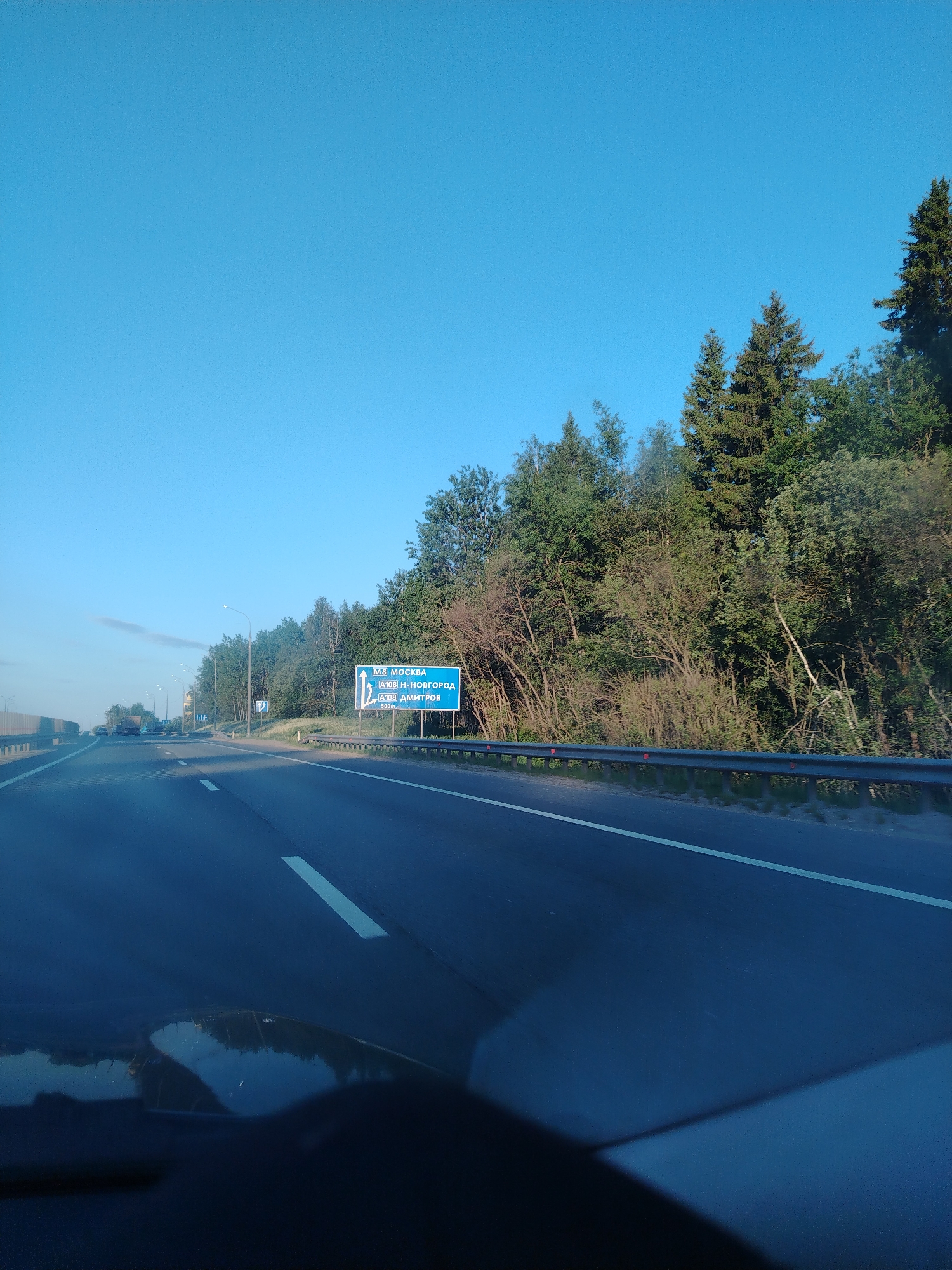 The image depicts a highway scene, likely taken from inside a vehicle in Bereznyaki, Russia. The road is asphalt with white lane markings. A blue road sign is visible, indicating directions to Moscow (M8), N-Novgorod (A108), and Dmitrov (A108). Trees line the side of the highway, and a metal guardrail separates the road from the vegetation. The sky is clear and blue. The vehicle's dashboard is partially visible in the foreground.