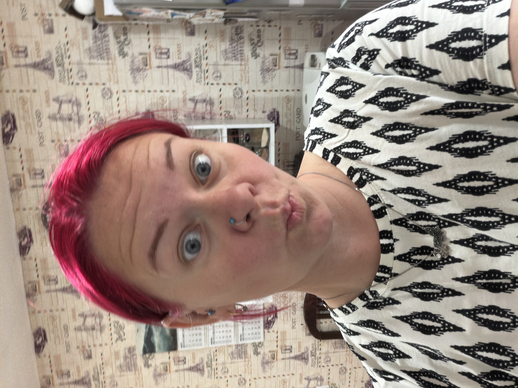 A woman with bright pink hair and blue eyes is taking a selfie. She is wearing a black and white patterned shirt and a silver necklace with a charm. She has a nose ring and earrings. The background is a wall with a "Post Card" pattern featuring images of the Eiffel Tower and other city landmarks. The woman appears to be indoors, possibly in a home. The specific location in Chaikovsky, Russia, is not identifiable from the visual information provided.