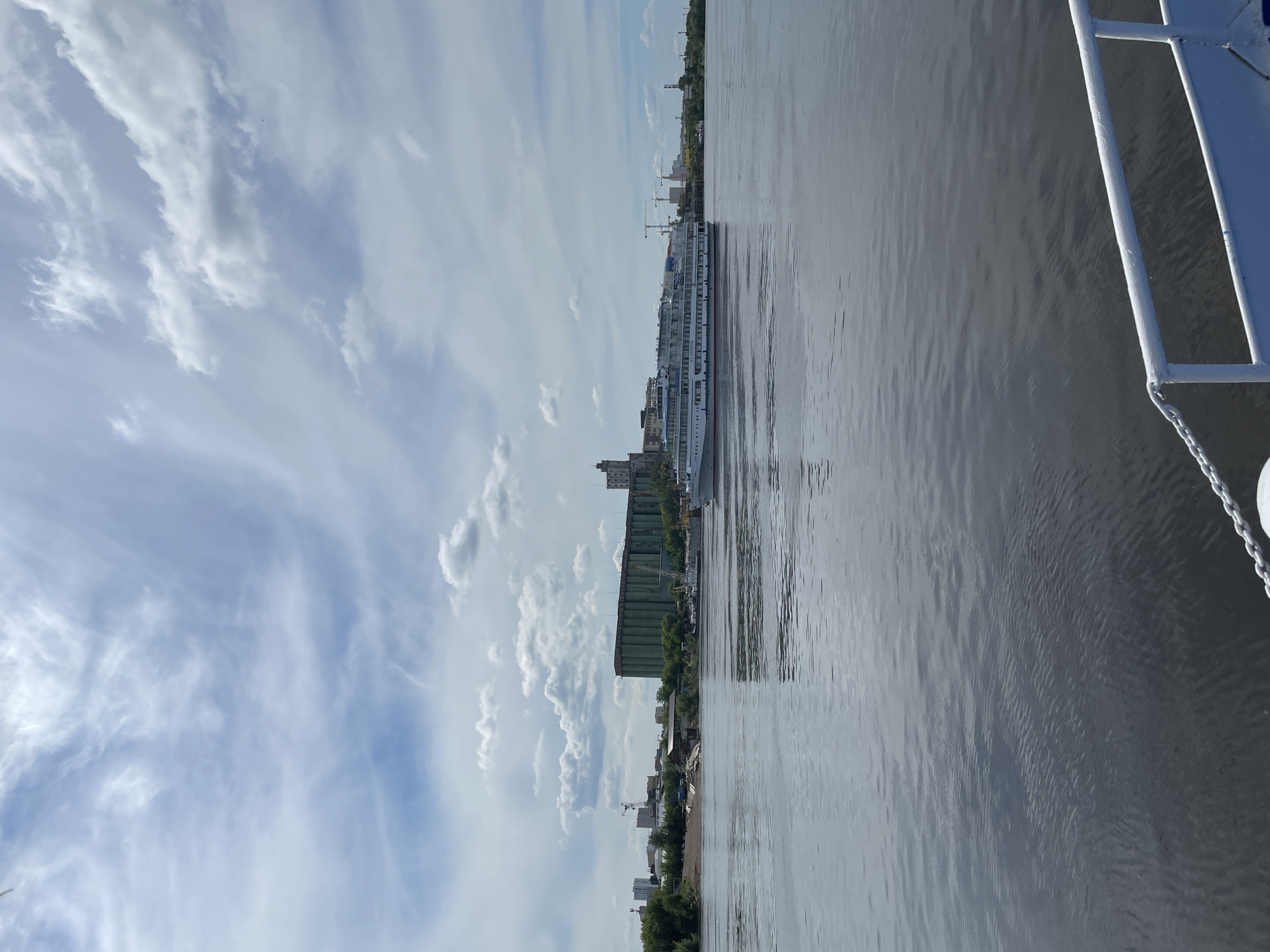 The media file captures a serene daytime river scene, presented from a perspective on the water. The original image is oriented vertically, but when viewed horizontally, it displays a wide river stretching across the middle ground. On the right side, a white boat railing and chain are partially visible, suggesting the photograph was taken from another vessel or a dock.
A prominent feature is a large, multi-deck white river cruise ship, docked along the left bank of the river. Its multiple levels and rows of windows are clearly discernible, indicating a significant passenger capacity. The river's surface is calm, with gentle ripples reflecting the soft daylight. The presence of the docked ship suggests either an arrival, departure, or a stopover during a journey.
The left bank of the river is characterized by an urban landscape. A mix of buildings, including residential and industrial structures, lines the shore, interspersed with green trees and foliage. In the distance, several construction cranes rise above the skyline, pointing to ongoing development.
The sky occupies the upper portion of the frame, showcasing a mix of blue patches and scattered white clouds. These range from elongated, wispy formations to more compact, fluffy cumulus clouds, all indicative of fair weather. The bright, diffused light suggests it is daytime, likely late morning or early afternoon. The overall atmosphere conveys tranquility and a sense of routine activity on the river. No visible text is legible on any objects within the image. Given the provided context, the scene is situated in Perm, Russia, likely along the Kama River.