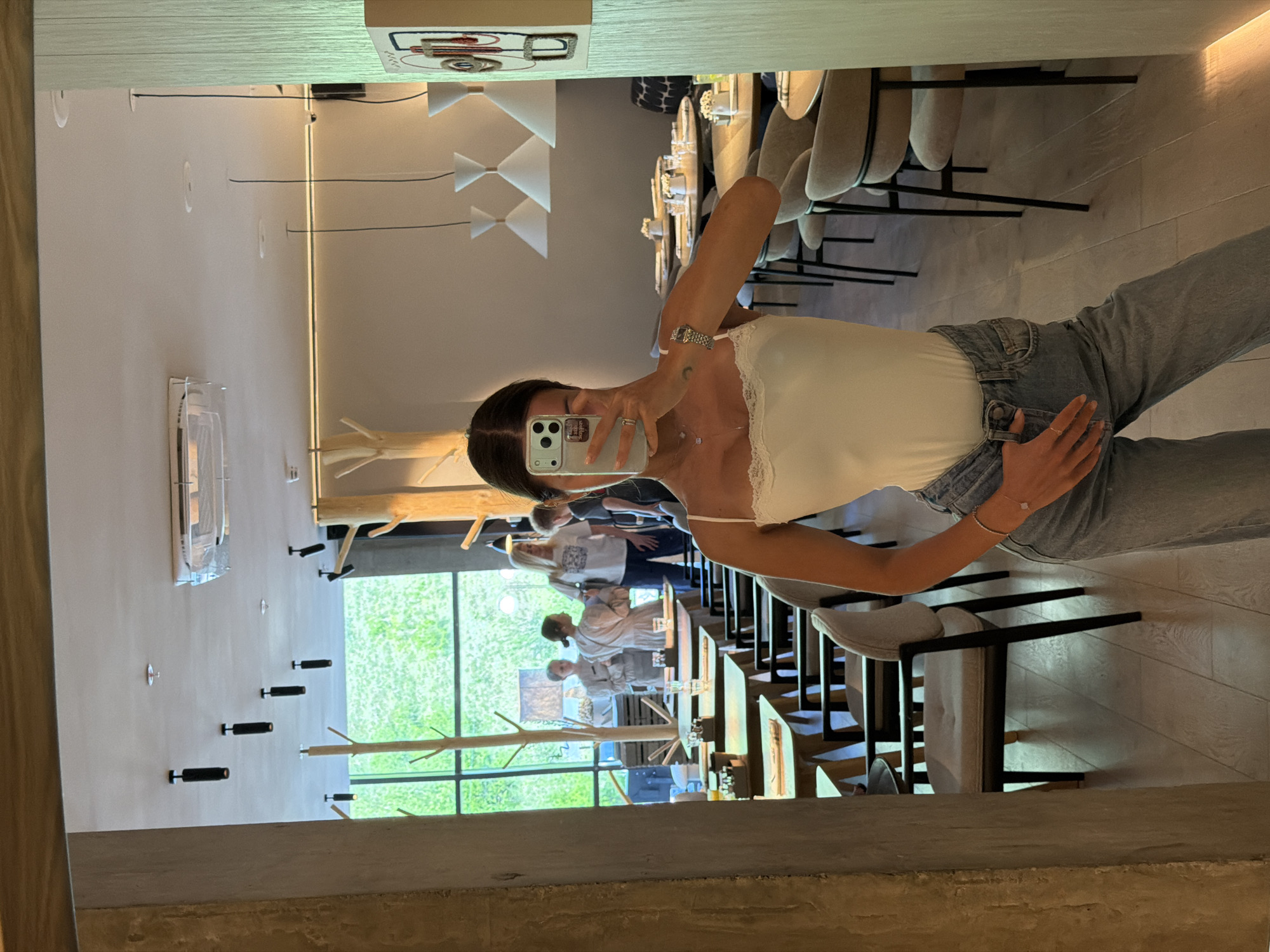 The image shows a reflection of a woman taking a selfie in what appears to be a restaurant. The restaurant has a minimalist and modern aesthetic, with light-colored walls, wooden accents, and contemporary lighting fixtures. In the reflection, the woman is wearing a white top and blue jeans. There are other people visible in the background through a large window, and tables are set up for dining, suggesting it is a restaurant or cafe. Based on the information provided, the location is Krasnoyarsk, Russia.