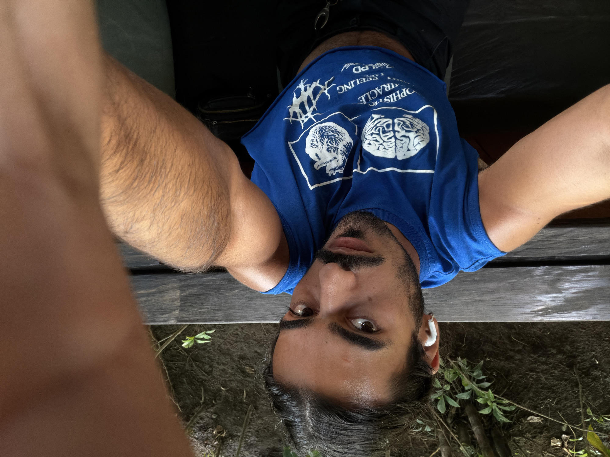 A medium shot captures a person from the chest up, wearing a blue sleeveless shirt with graphic prints of a brain and a heart. The person has dark hair pulled back and is wearing white earbuds. They have dark eyes, a mustache, and stubble. The person is holding a phone out of frame, suggesting they are taking a selfie. They are sitting on a wooden bench outdoors, with lush green foliage and trees visible in the background. The setting appears to be a park or garden area. The lighting suggests it is daytime.
