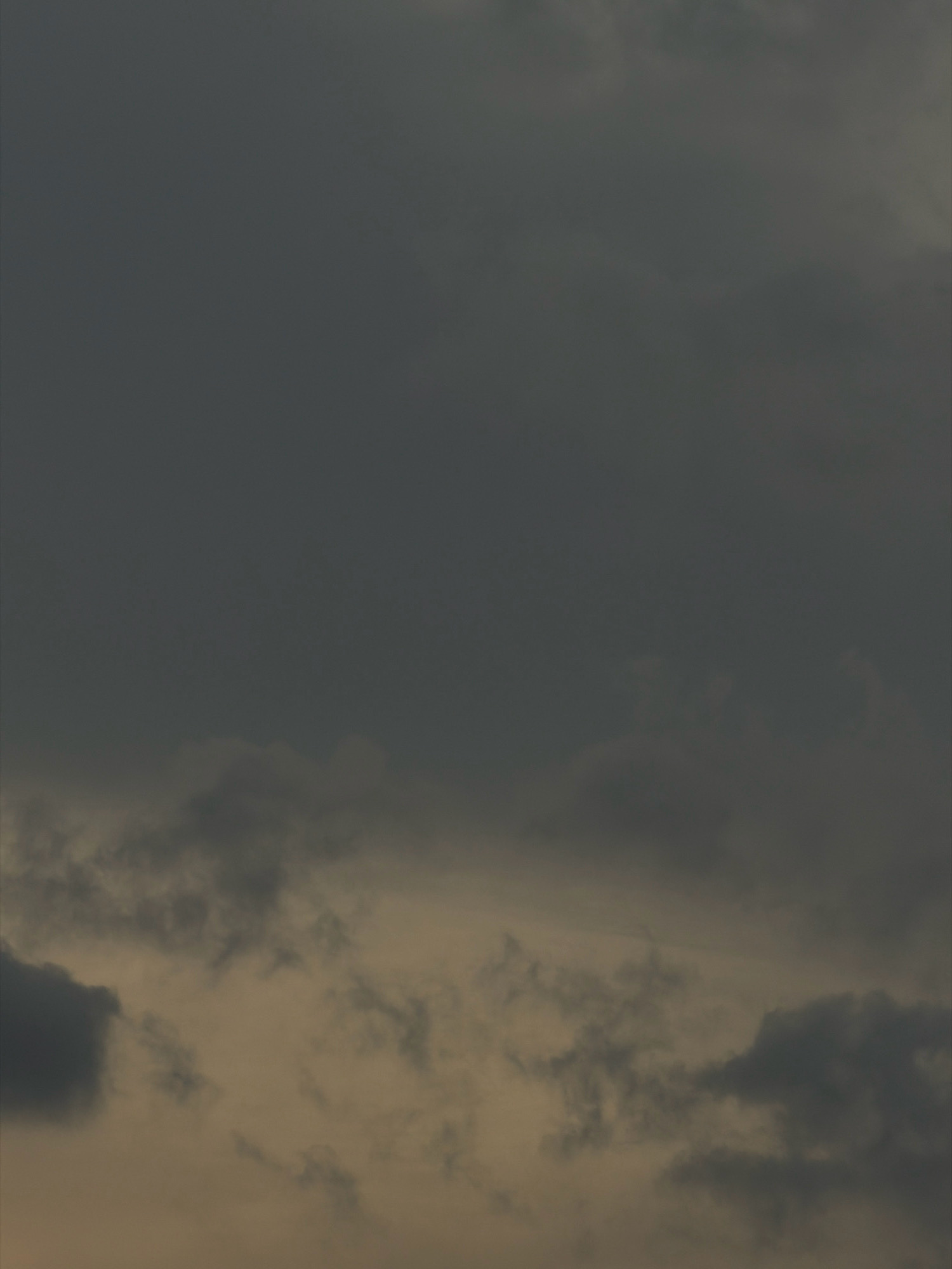 The image depicts a cloudy sky over Koorkaparamba, India. The upper portion of the frame is dominated by a dark, overcast sky, suggesting potentially inclement weather or the approach of evening. The lower portion of the image features lighter, more diffused clouds with a yellowish or brownish hue, possibly indicating scattered sunlight breaking through or a layer of haze. There are no visible people, structures, or specific landmarks that would allow for a more precise identification of the location within Koorkaparamba. The scene is purely atmospheric, focusing on the cloud formations and the overall mood conveyed by the lighting.