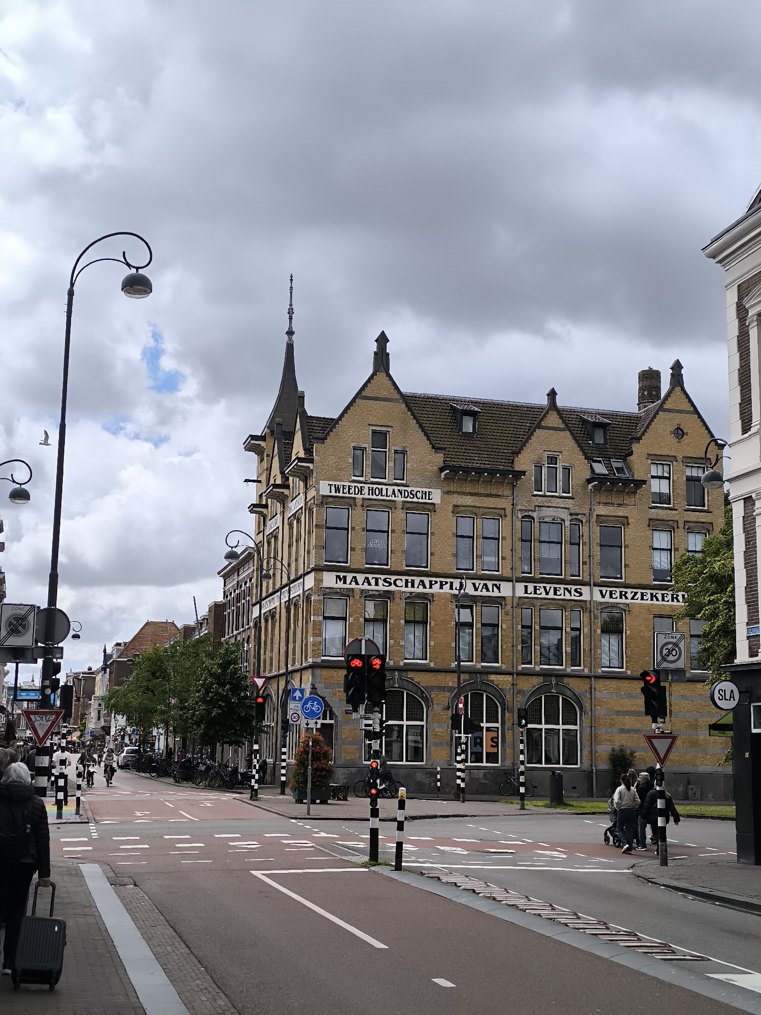 The image captures a street scene in Haarlem, Netherlands, focusing on a yellow brick building with the text "TWEEDE HOLLANDSCHE MAATSCHAPPIJ VAN LEVENS VERZEKERINGEN" visible on its facade. The building exhibits a traditional architectural style with multiple windows and decorative elements. In the foreground, a street with pedestrian crossings and traffic lights is visible. Several people are present, some walking with luggage, others crossing the street. Bicycles are also visible, parked along the street and being ridden in the distance. The sky is overcast with clouds. The overall environment suggests an urban setting with a mix of historical architecture and modern traffic infrastructure.