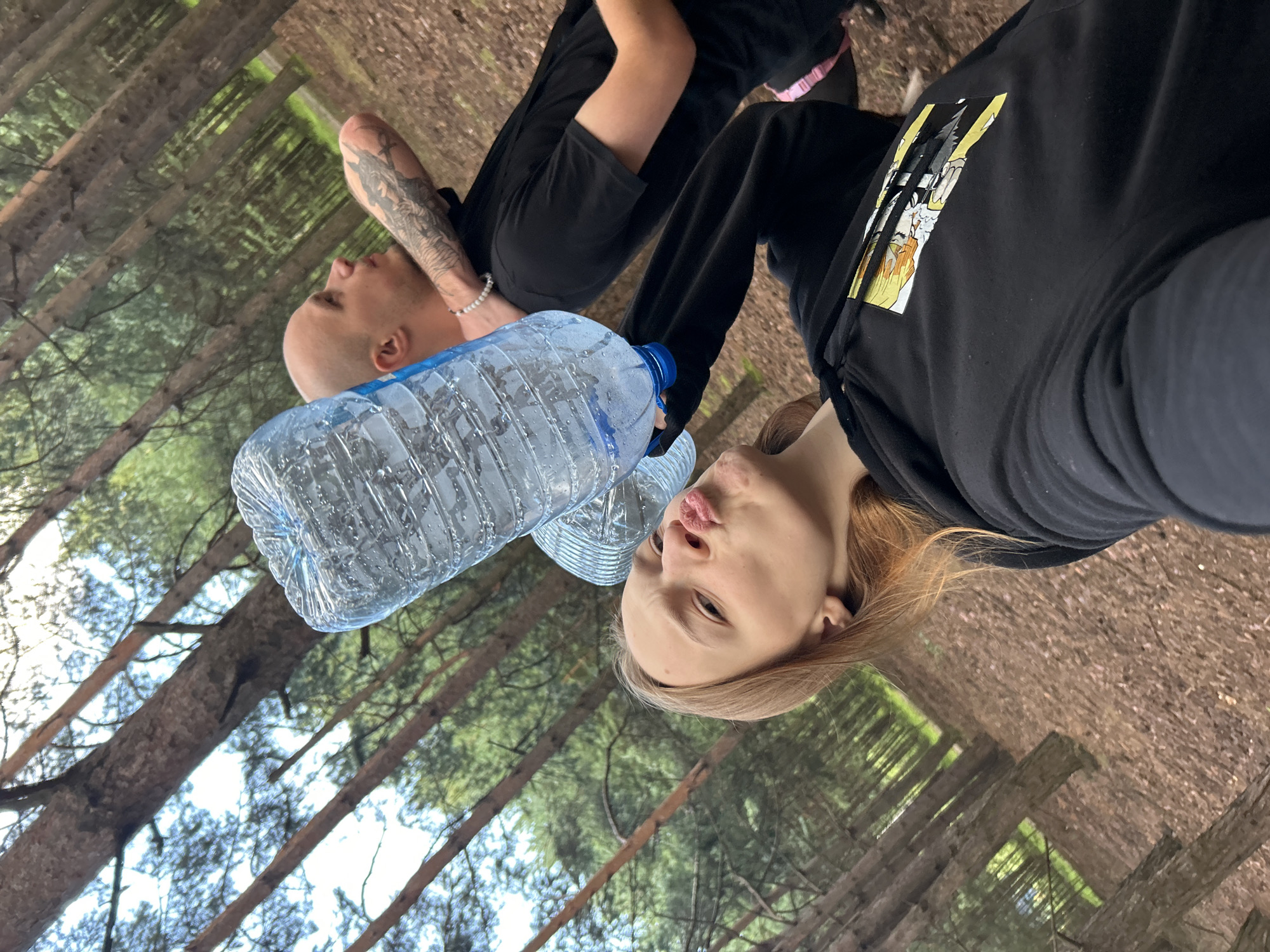 In an outdoor, wooded environment consistent with a park or forest, two individuals are present. One individual, a bald male with a tattoo on his arm, is positioned in the background, looking to the right. He is wearing a black t-shirt. In the foreground, a female with blonde hair is holding two large plastic water bottles. She is looking at the camera with her lips pursed. She is wearing a black hoodie. The ground is covered with pine needles, and pine trees are visible in the background. The lighting suggests daytime. The image appears to be taken in the city of Chaikovsky, Russia, based on the provided context.