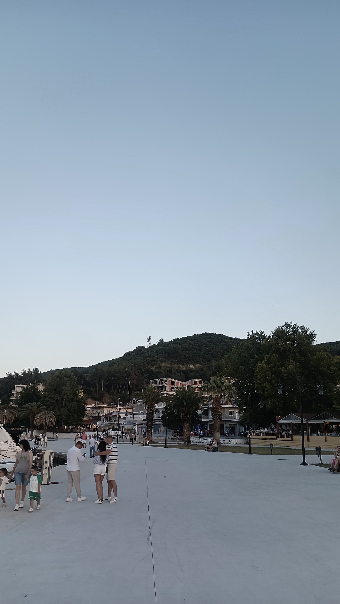 The image depicts a wide, paved public area in Νέα Ηρακλείτσα, Greece, likely a waterfront promenade or plaza. A clear, pale blue sky dominates the upper portion of the frame. In the background, a lush green hillside rises, dotted with buildings and topped by a prominent telecommunications tower. Palm trees and other foliage are visible along the base of the hill, interspersed with more modern buildings, some appearing to be under construction.
Several groups of people are scattered across the paved area. On the left, a family consisting of two adults and two children are walking, with a woman in a grey t-shirt and shorts, a man in a white t-shirt and beige pants, and two young children. To their right, a couple stands closely together, with the woman in a striped t-shirt and white shorts, and the man in a white t-shirt and white shorts. Another man in a white shirt and light-colored pants stands nearby. Further into the background, more people are visible, some seated at outdoor tables of establishments that appear to be restaurants or cafes. The lighting suggests it is late afternoon or early evening, with the sun low on the horizon.
