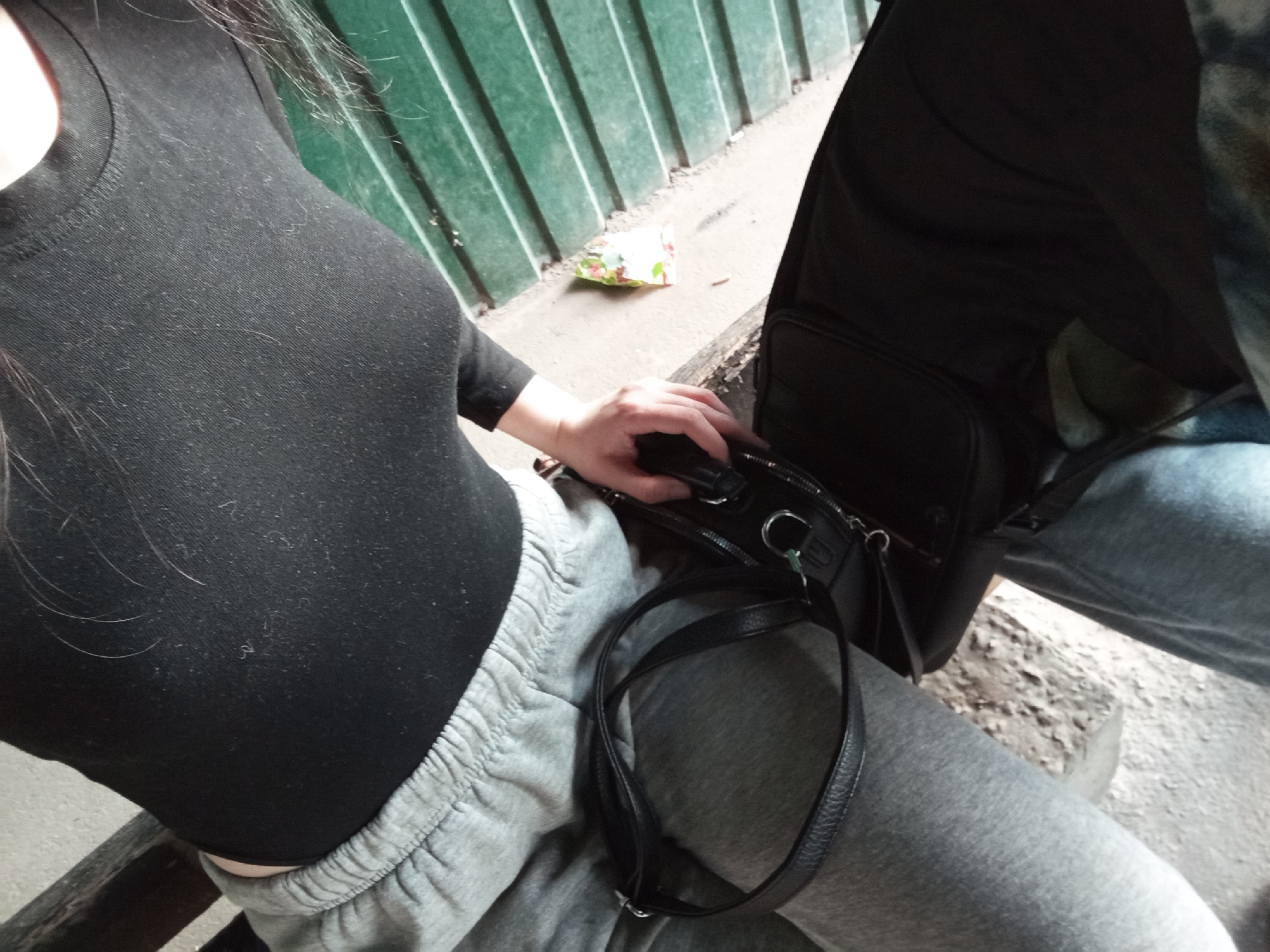 The image shows a close-up, high-angle view of two people sitting outdoors, likely in Poltava, Ukraine. The primary subject on the left is seen from the chest down, wearing a form-fitting black long-sleeved top and grey sweatpants with an elasticated waistband. Their dark hair is visible over their left shoulder. Their right hand rests on a black handbag with a strap and silver hardware, which is placed on their lap or between the two individuals.
To the right, a second person is partially visible, wearing blue jeans and an upper garment with a camouflage or patterned design. A black backpack or large bag is also seen next to them.
The setting features a dark green corrugated metal fence or wall in the background. The ground is a light-colored concrete or asphalt surface with some dirt and debris. A small, crumpled piece of litter, possibly a food wrapper with green, white, and red colors, lies on the ground near the fence.
The activity depicted is simply two people sitting together. The time of day is daylight, but specific weather conditions or emotions are not discernible. The black top of the person on the left has visible lint or fibers on its surface. There is no visible text in the image.