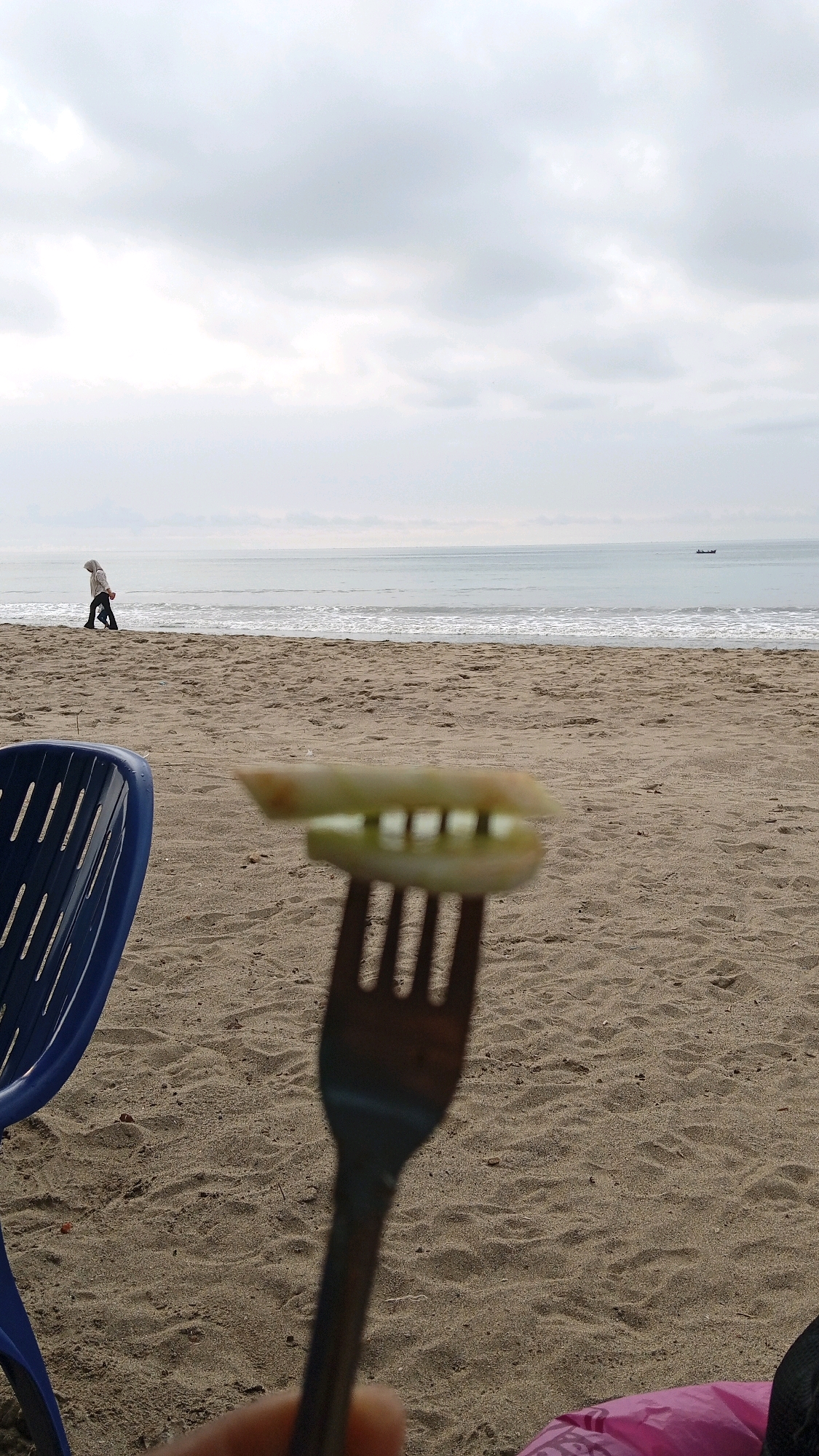 A person walks on a sandy beach towards the ocean under an overcast sky. The water is calm with small waves. In the foreground, a fork holds a slice of cucumber. A blue plastic chair is visible on the left side of the frame. The scene is likely at Kruengjuli Timu, Indonesia, given the context.