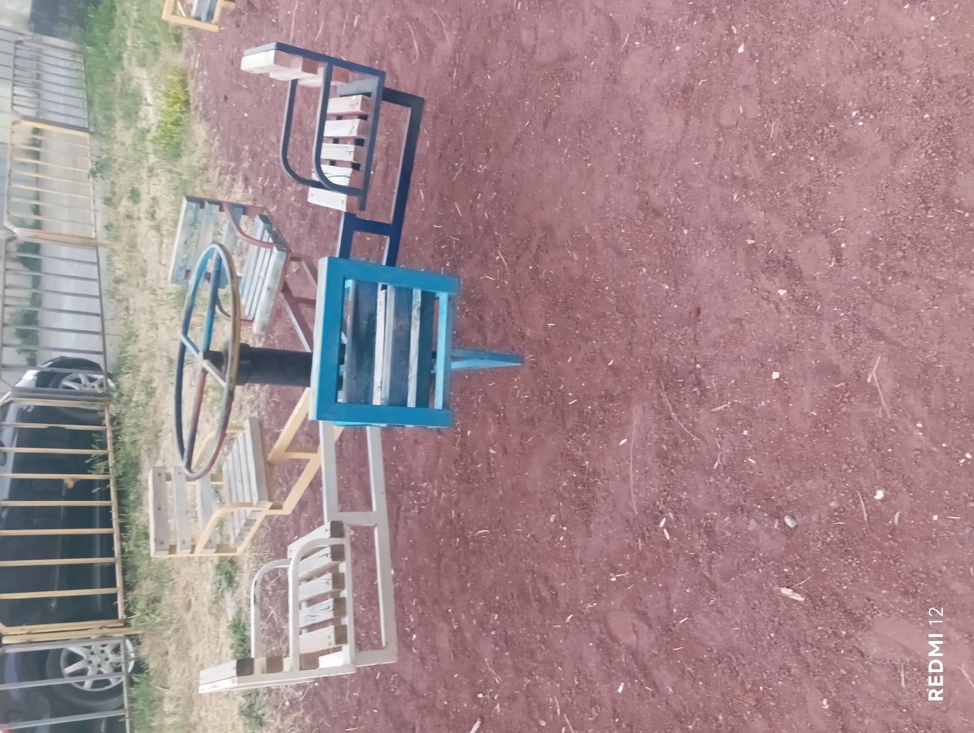 This media file depicts a playground merry-go-round with multiple seats, set on a dirt or gravel surface. The merry-go-round has a metal frame with wooden slats forming the seats. One seat is painted blue, while others are natural wood or possibly painted white. A blue-rimmed steering wheel is visible in the center. To the left of the merry-go-round, a black vehicle is partially visible, with its tires and a portion of its side showing. Behind the vehicle is a fence made of yellow vertical bars. The ground is covered in reddish-brown dirt with some dry grass and scattered debris. The lighting suggests it is daytime, possibly late afternoon, with no significant shadows indicating harsh midday sun. The scene appears to be outdoors in an open area, likely a park or recreational space. There is no discernible activity taking place, as the merry-go-round is stationary and no people are present. Visible text in the bottom right corner reads "REDMI 12".