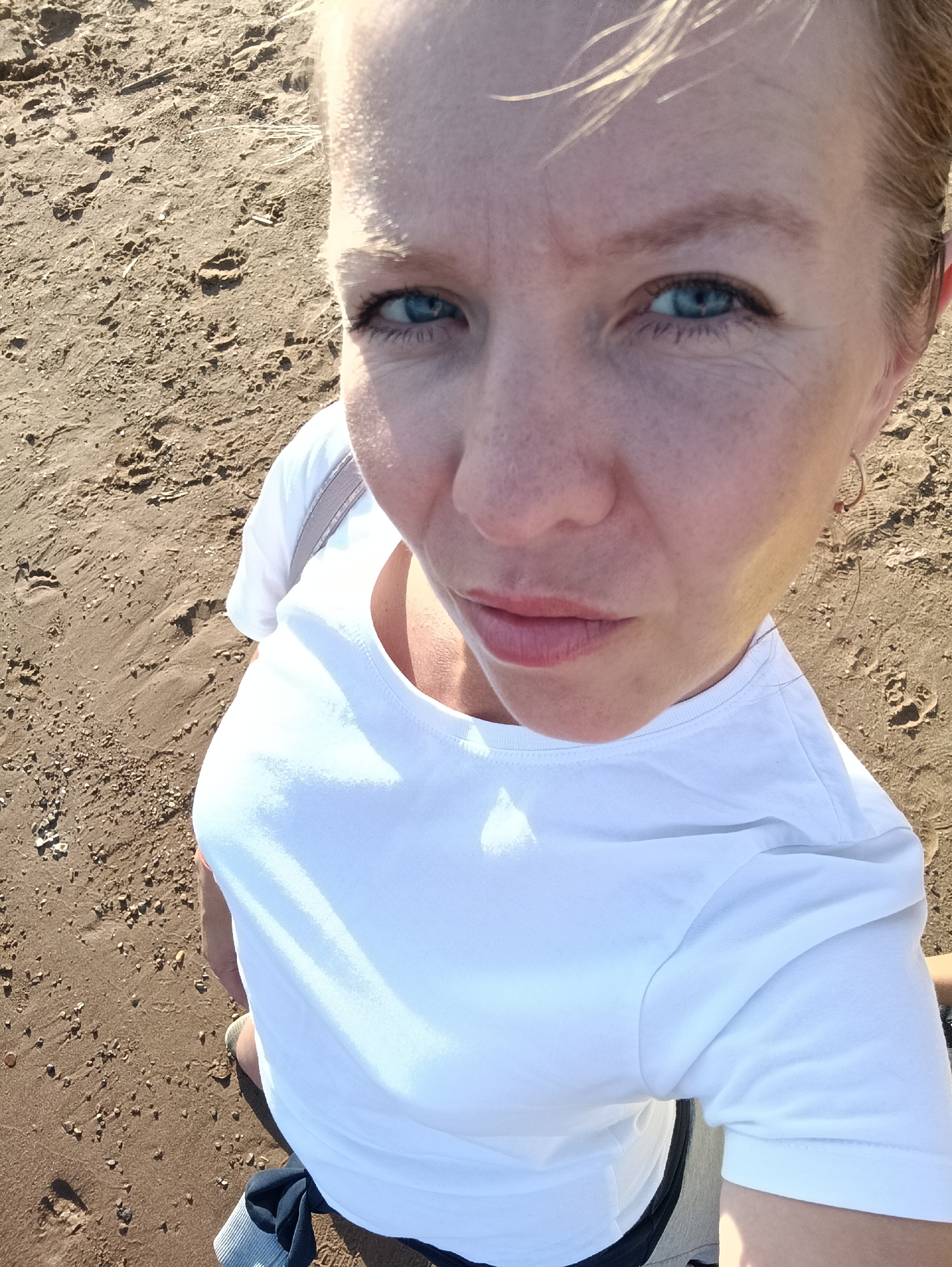 This is a high-angle, close-up selfie of a woman standing on a sandy beach. She has short blonde hair and blue eyes, and she is wearing a white t-shirt. The sand is a light brown color with small pebbles and footprints visible. The lighting suggests it is daytime, likely sunny. Her expression is a bit ambiguous, perhaps curious or thoughtful. The overall impression is of a casual moment captured outdoors. There is no visible text in the image.