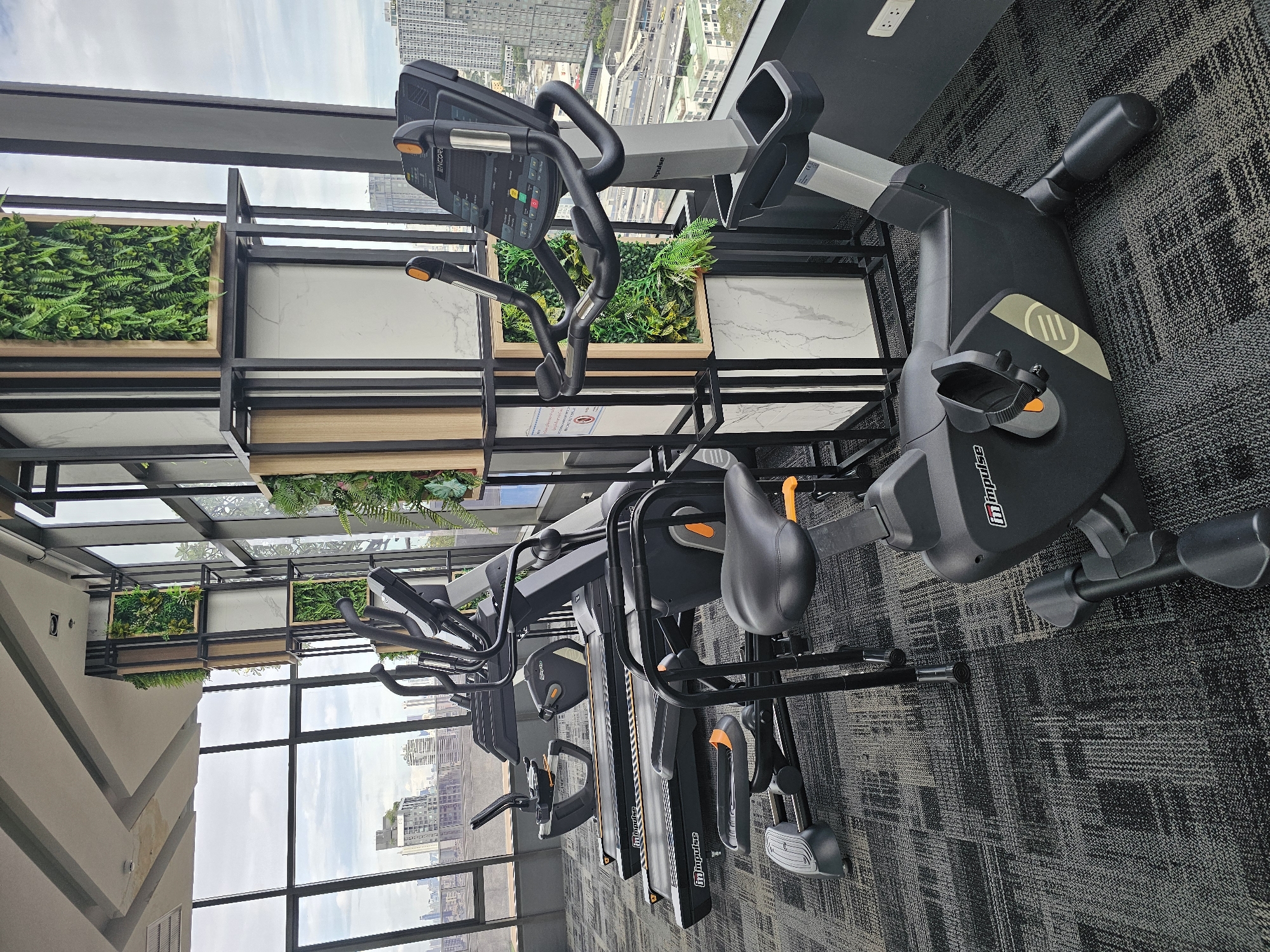 A gym is situated on a high floor in a city, likely Bangkok, Thailand, as evidenced by the dense urban skyline visible through the large windows. The gym is equipped with several pieces of cardio equipment, including treadmills and an exercise bike, all arranged on a patterned carpet. The exercise bike in the foreground has a gray and black frame with orange accents and a digital display panel. Behind it, three treadmills are lined up, each with the "Impulse" logo visible. The gym features a modern interior design with black metal shelving units containing artificial greenery and marble-patterned panels. The overall environment appears clean and well-maintained, offering a view of the city from its elevated position.