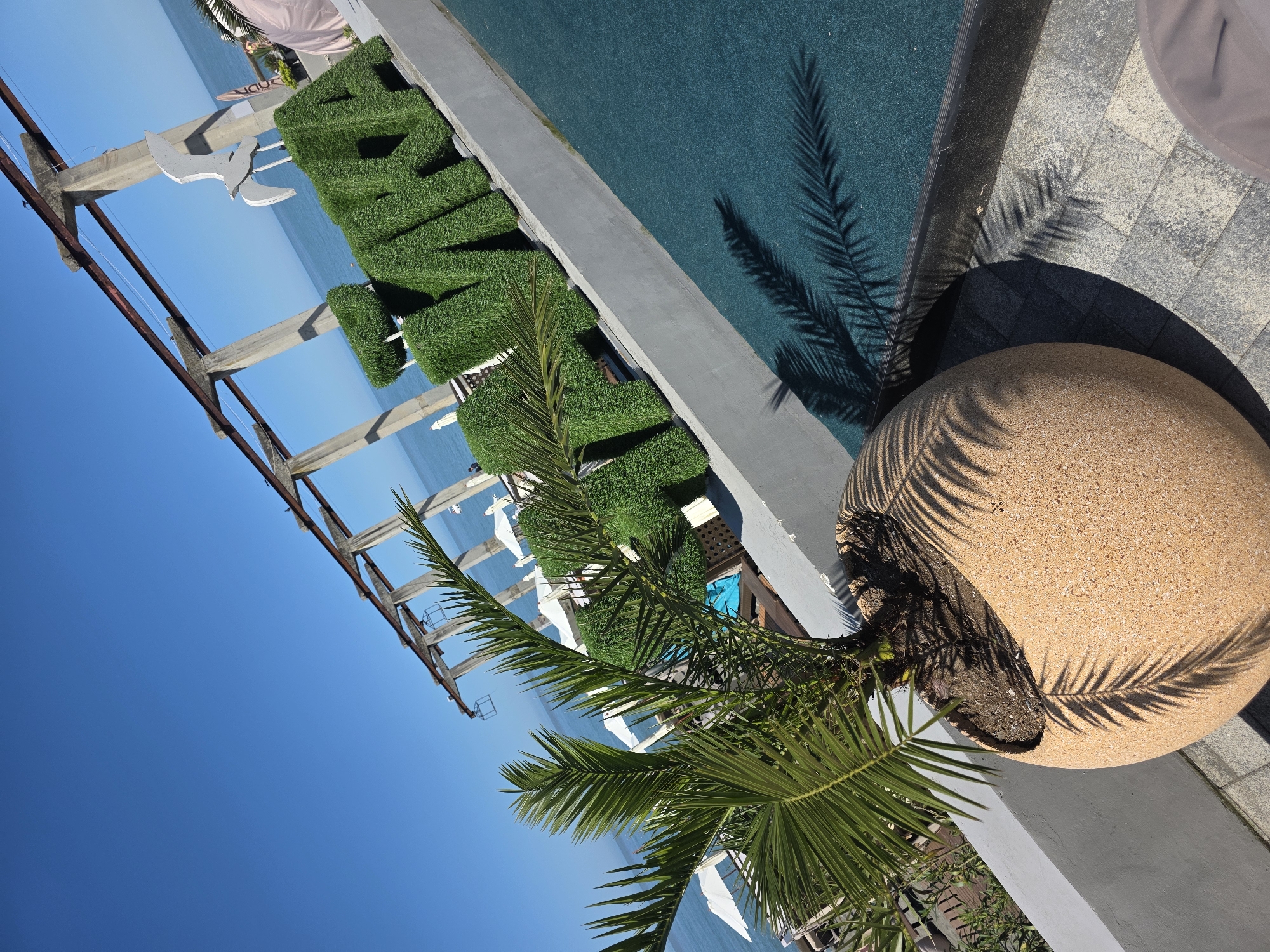 The image depicts a bright, sunny coastal scene in Sochi, Russia, observed from a slightly elevated and tilted perspective. A clear, deep blue sky meets a calm, light blue sea at the horizon.
In the immediate foreground, a small, vibrant green palm tree stands in a large, spherical, light-brown textured planter. Distinct, sharp shadows of the palm fronds are cast onto the planter, the grey interlocking paver stones, and a section of teal-colored outdoor carpet or artificial turf on the ground. A rounded, light-colored object, possibly a hat or part of a chair, is partially visible in the bottom right corner.
Behind the foreground area, a low, light-grey concrete retaining wall runs diagonally across the frame. On the far side of this wall, large, three-dimensional letters covered in artificial green turf spell out "РИВЬЕРА" (Riviera). Positioned behind and above the "РИВЬЕРА" sign is a white cutout of a seagull in flight.
Further into the midground, beyond the sign, multiple white sun umbrellas and blue and white sun loungers are visible on what appears to be a terraced area or a beach adjacent to the sea. Two indistinct human figures are faintly discernible under two of the white umbrellas in the distance.
Spanning the upper midground and background is a large, weathered structure composed of concrete pillars and horizontal metal beams, possibly remnants of an old pier, an industrial frame, or a pergola, extending out towards the sea. The strong sunlight illuminates the scene, creating prominent shadows.