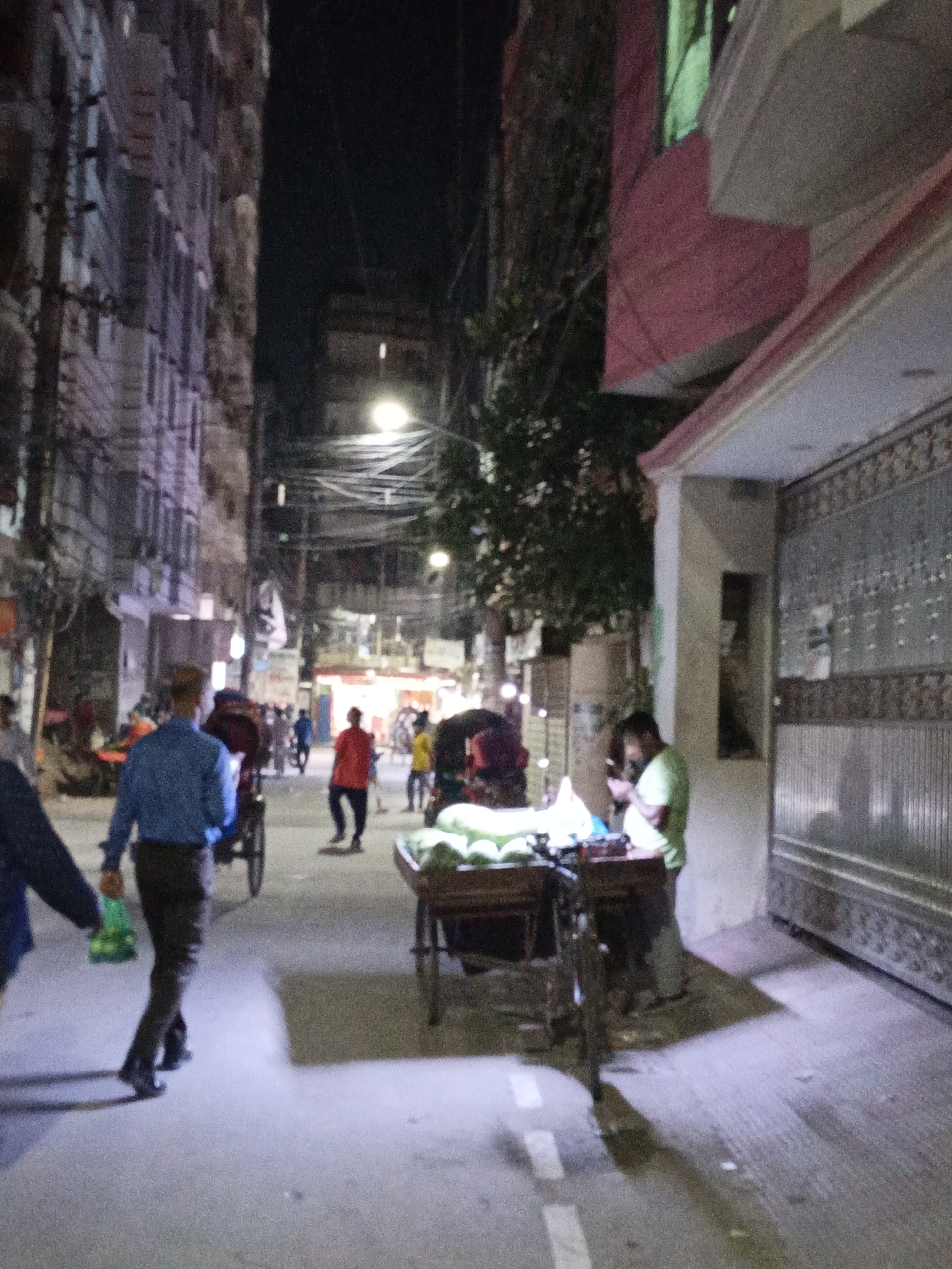 A dimly lit street scene in Dhaka, Bangladesh. People are walking on the street. A man in a blue shirt is walking away from the camera. A man stands at a cart. A red-shirted man is walking toward the center of the frame. Other people are visible in the background, along with rickshaws. Overhead are many electrical wires, and streetlights. Buildings are on both sides of the street. The house on the right has a large gate.
