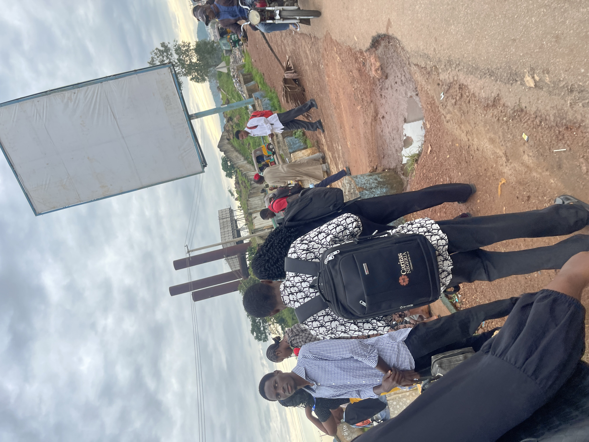 In Rafin Par, Nigeria, under a cloudy sky, a group of people stand on a dirt road beside a paved area. The road is uneven and muddy, with puddles of water. In the background, several tall, cylindrical structures, possibly industrial smokestacks, rise into the sky, along with power lines. A large, blank billboard on a pole is tilted at an angle. Several individuals, including young men and women, are visible. One young man in the foreground wears a checkered blue and white shirt and black pants, with his hands clasped in front of him. Another young man behind him wears a white shirt with a black pattern and a black backpack with the "Caritas Nigeria" logo. A man in a blue jacket rides a motorcycle in the background. Other people are seen in the middle ground, some walking or standing. Small vehicles, including what appears to be a tricycle taxi, are also present. The scene suggests a public area, possibly a street or gathering point, with evidence of a transitional or developing environment.