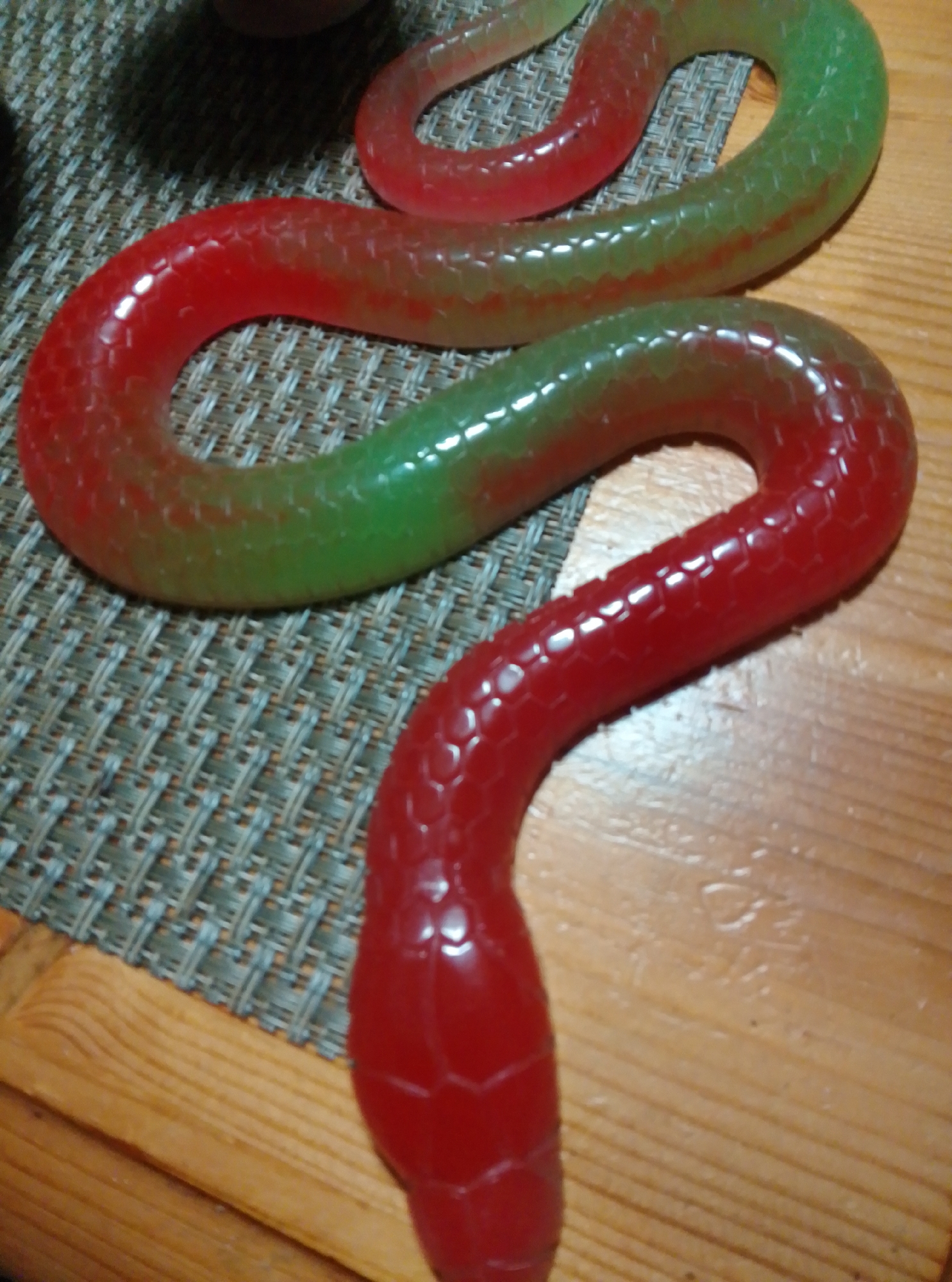 A gummy candy in the shape of a snake lies on a woven placemat and a wooden surface. The candy transitions in color from red at the head to green at the tail. The texture of the candy mimics snake scales. The setting does not provide any information about the city of Wedel, Germany.