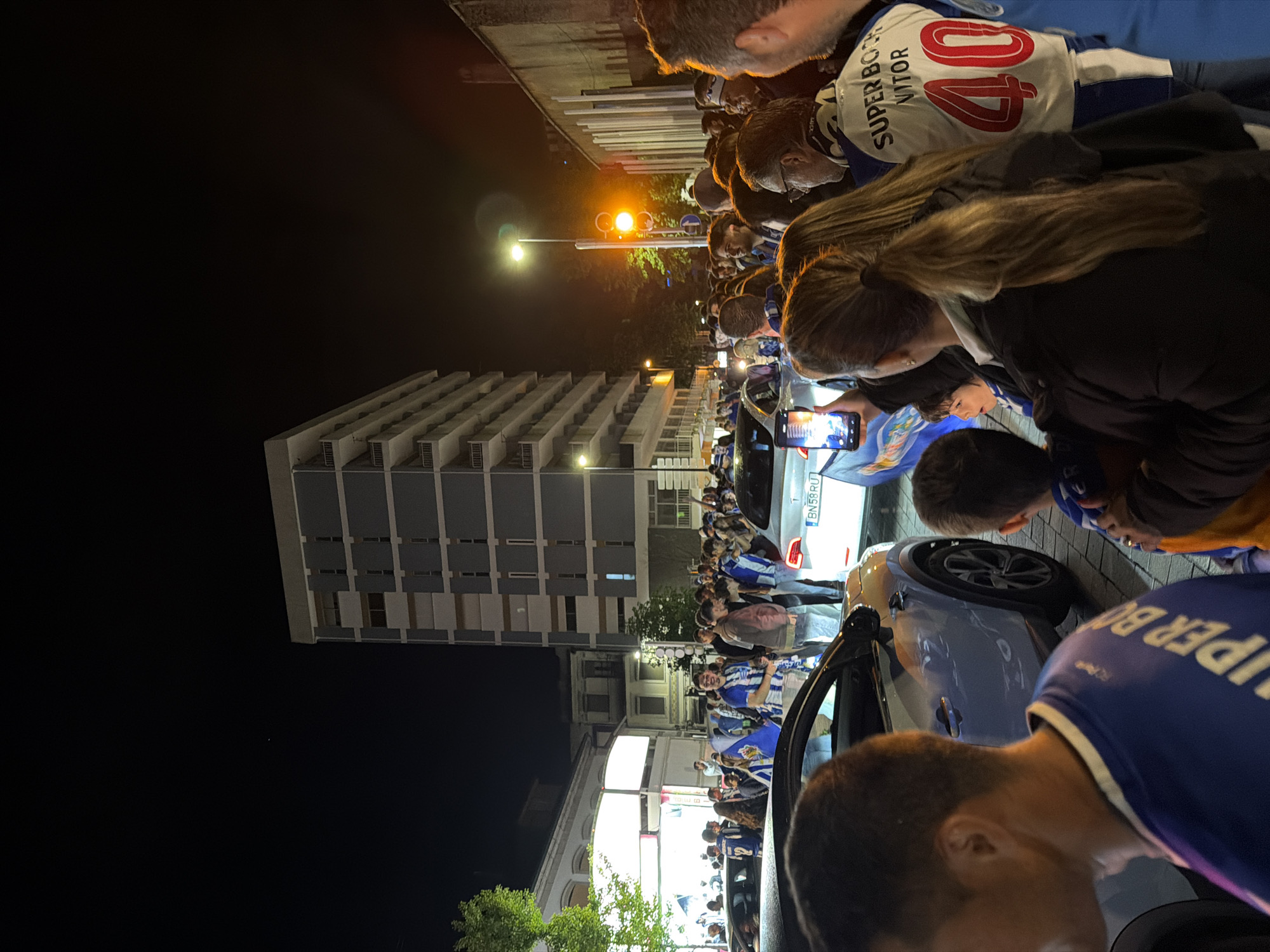 At night in Vila Real, Portugal, a crowd of people is gathered along a street, likely celebrating. They are wearing blue and white clothing and scarves, with some items labeled "SUPER BOCK VITOR 40." A light blue car is parked on the street with the Portuguese license plate "BN59RU". A tall, modern apartment building with multiple floors is visible in the background. A street lamp is illuminated. One person is holding up a smartphone to record or photograph the scene.