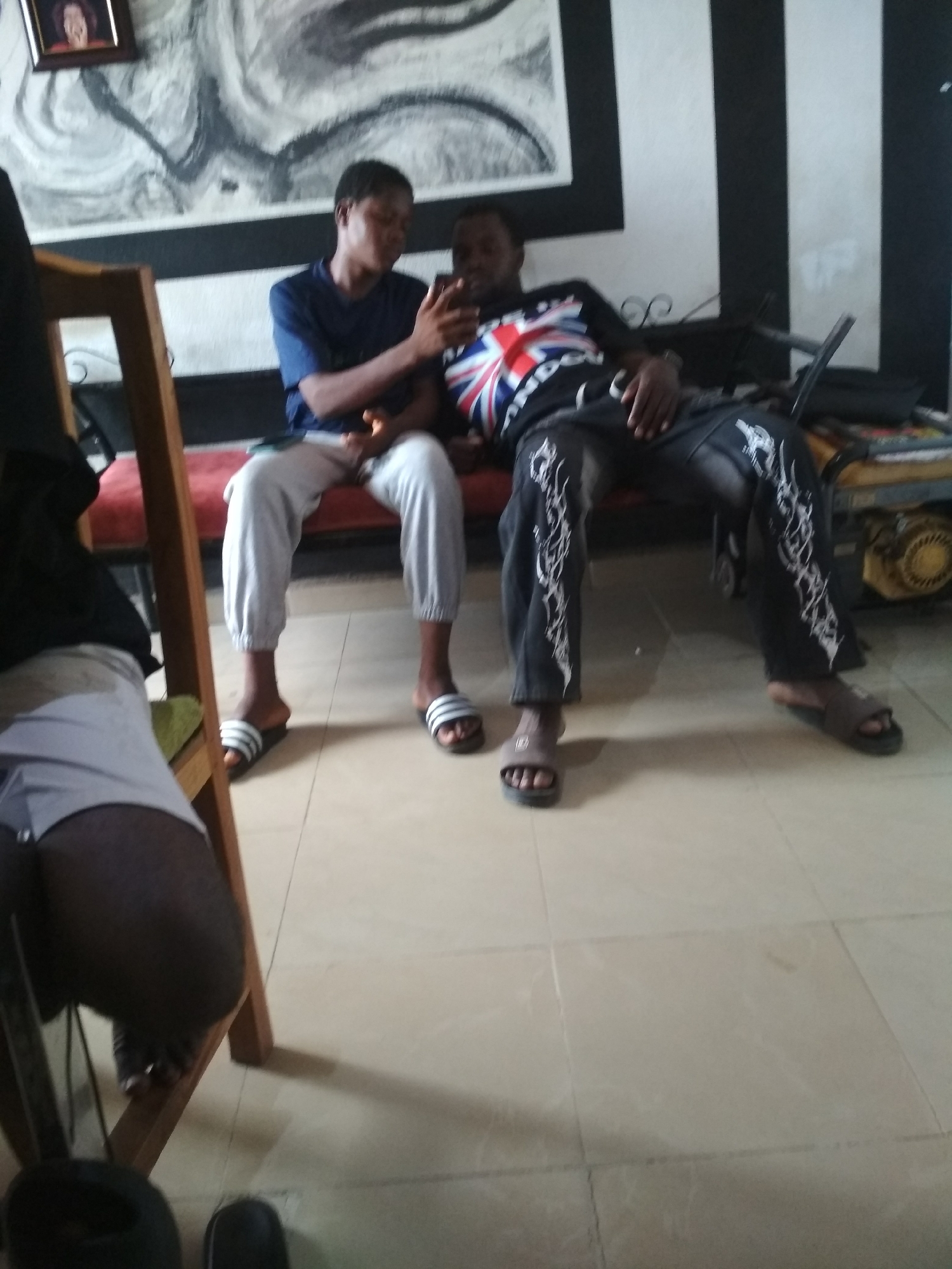 Here is a detailed description of the image:
**Scene Overview:** The image appears to be taken indoors, likely in a residential space, given the presence of furniture and decor. The lighting is moderately bright, suggesting daytime.
**People:**
* Two young men are seated together on a red cushioned bench. One is wearing a blue shirt and gray sweatpants, while the other wears a dark shirt with a Union Jack (British flag) design, along with patterned dark pants.
* The man in the blue shirt is leaning towards the other, possibly showing him something on a mobile phone.
* Part of a third person is visible in the foreground to the left, showing a black shirt and white shorts.
**Objects:**
* Mobile phone being held by the man in the blue shirt.
* Red cushioned bench where the two men are sitting.
* Wooden chair to the left of the frame.
* An electrical generator is visible in the background.
* The wall features a black and white abstract artwork and vertical black and white stripes.
* A framed picture hangs on the wall.
* Sandals are worn by all three individuals.
**Actions:**
* The two men are interacting and focused on the mobile phone.
* The environment suggests a relaxed, casual setting.
**Interactions:**
* The man in blue appears to be showing something on the phone to the man in the British flag shirt.
**Environment:**
* The background has a simple yet slightly cluttered appearance.
* The floor has a light beige tiled surface.
**Location:** The precise address is unknown. However, based on the style of the residence, it is in the city of Unknown, Unknown.