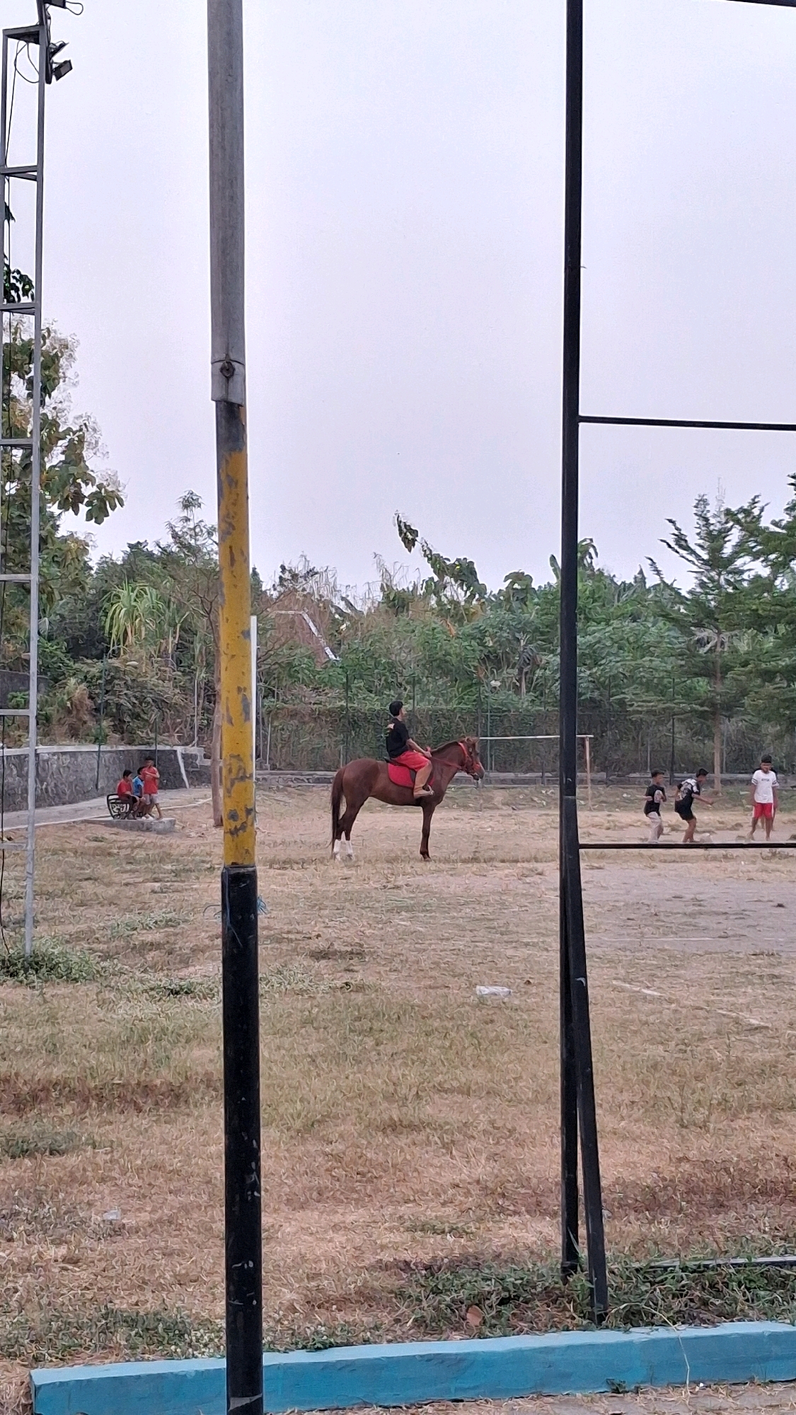 In an outdoor recreational area in Banguntapan, Indonesia, a brown horse with a rider wearing red shorts and a dark shirt stands on dry, grassy terrain. The rider appears to be a young male looking towards the right of the frame. In the background, a line of trees and foliage borders a fence. To the left, three young males are seated on a bench near a stone wall. To the right, a group of four young males are engaged in what appears to be a game of soccer or a similar sport. The scene is framed by tall, dark poles and a yellow and gray pole with a light fixture attached at the top. The sky is overcast.