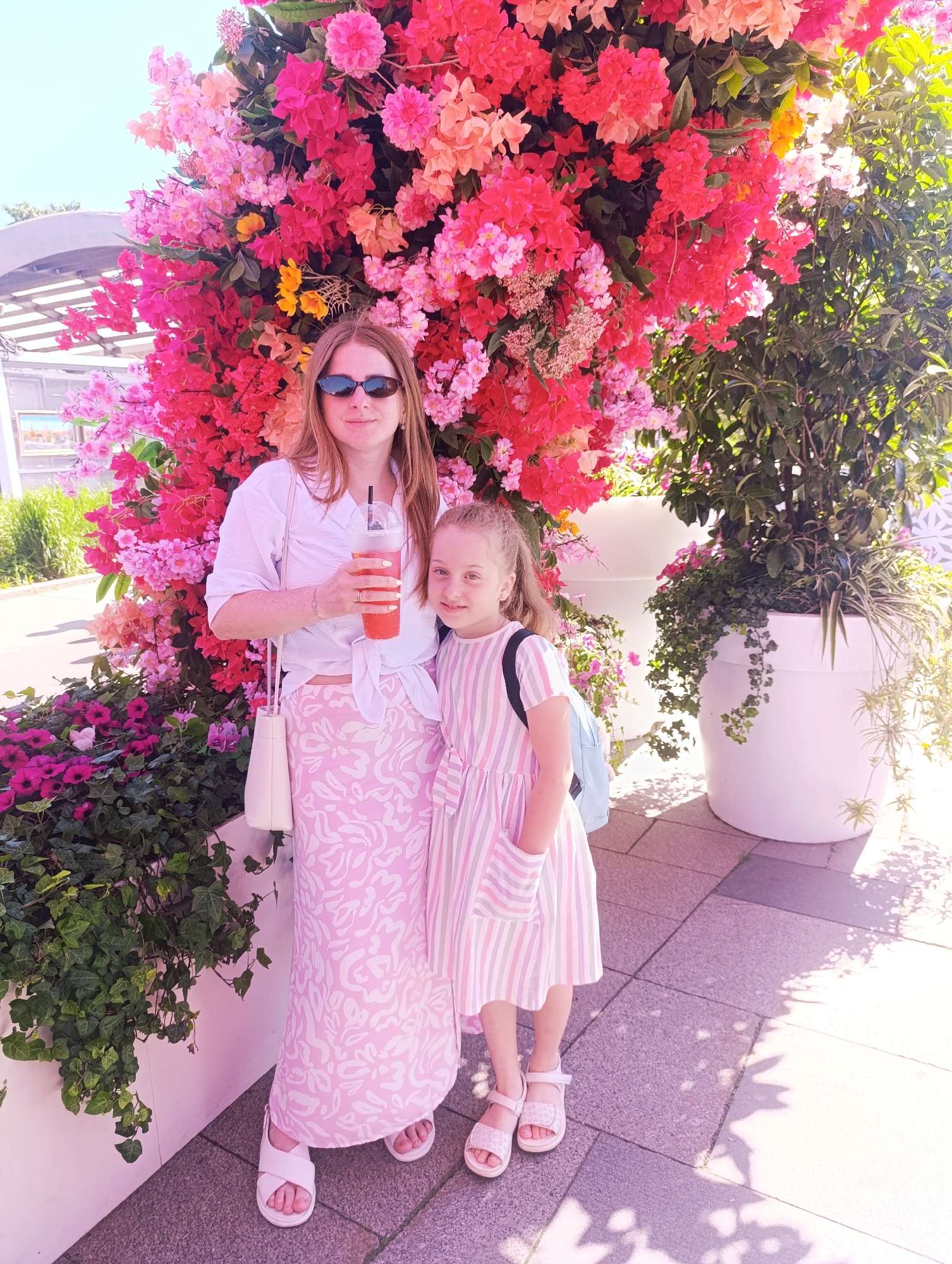 A candid outdoor portrait features two light-skinned females, an adult and a child, standing in full daylight in front of a prominent floral display. The adult female, positioned slightly to the left and behind the child, has long light brown hair, wears dark oval sunglasses, a white collared shirt tied at the waist revealing a sliver of midriff, and a long pink and white patterned skirt with white flat sandals. She holds a clear plastic cup with a reddish beverage and a black straw in her right hand; a small white shoulder bag is visible on her left. The child female, positioned to the right and slightly in front, has light brown hair pulled back into a ponytail and wears a pink and white striped sleeveless dress with pockets and white flat sandals. A light blue backpack with black straps is visible on her back. Both individuals face the camera with subtle smiles.
The dominant background is a dense, tall wall of artificial flowers, primarily in shades of vibrant pink, fuchsia, red, and orange, interspersed with some yellow and lighter pink blooms and green foliage, resembling bougainvillea. At the base of this floral wall, white rectangular planters contain dark green ivy and other plants. To the right of the child, two large, round, white planters hold tall, leafy green plants, some with cascading vines. The ground is paved with dark grey square or rectangular tiles, displaying dappled shadows from unseen overhead foliage, indicating direct sunlight. In the far left background, a curved, light-colored architectural structure with visible beams is seen beneath a clear light blue sky. A small framed picture depicting a cityscape is visible on a stand further left. The far right background reveals more dense green foliage and a light-colored wall with a subtle geometric pattern.