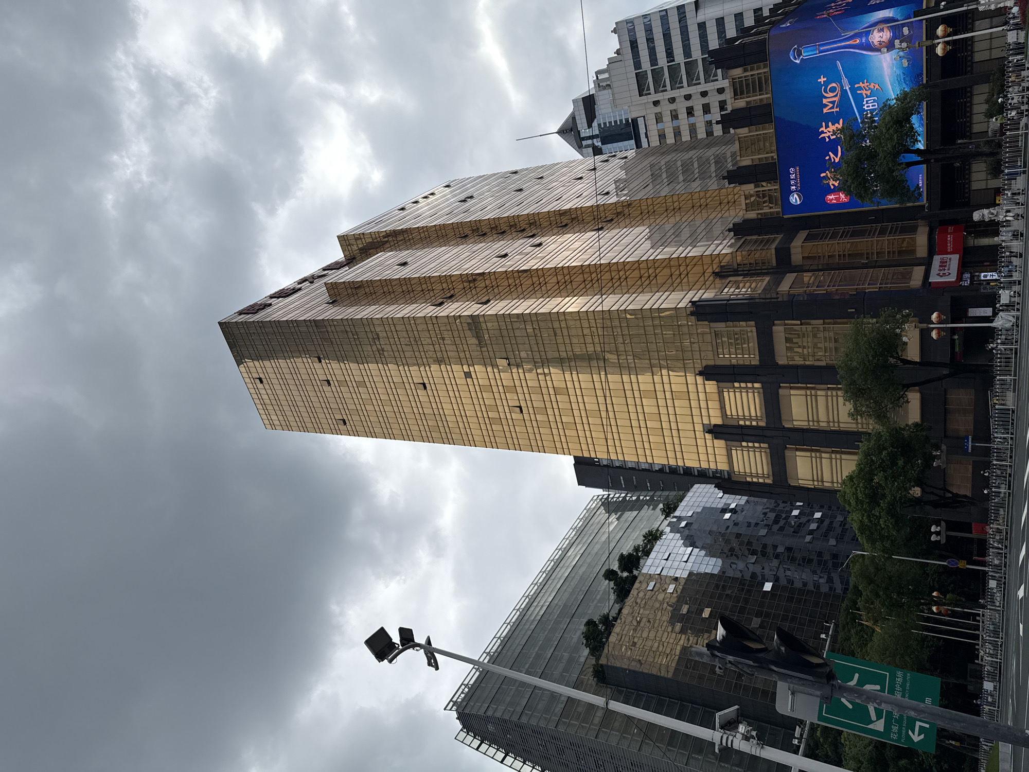 The image, taken in Tianhe District, China, depicts a street scene dominated by modern architecture under an overcast sky. A prominent golden skyscraper, likely a commercial building, is a central feature, its facade reflecting the muted light. To its left, a contemporary glass building rises, with intricate patterns on its exterior. In the foreground, a traffic light stands tall next to a green directional sign with Chinese characters and an arrow pointing left. The street is lined with a metal fence and trees, with a few people visible on the sidewalk. A large billboard displays a vibrant advertisement, adding a splash of color to the otherwise muted urban landscape. The overall impression is one of a bustling commercial district.
