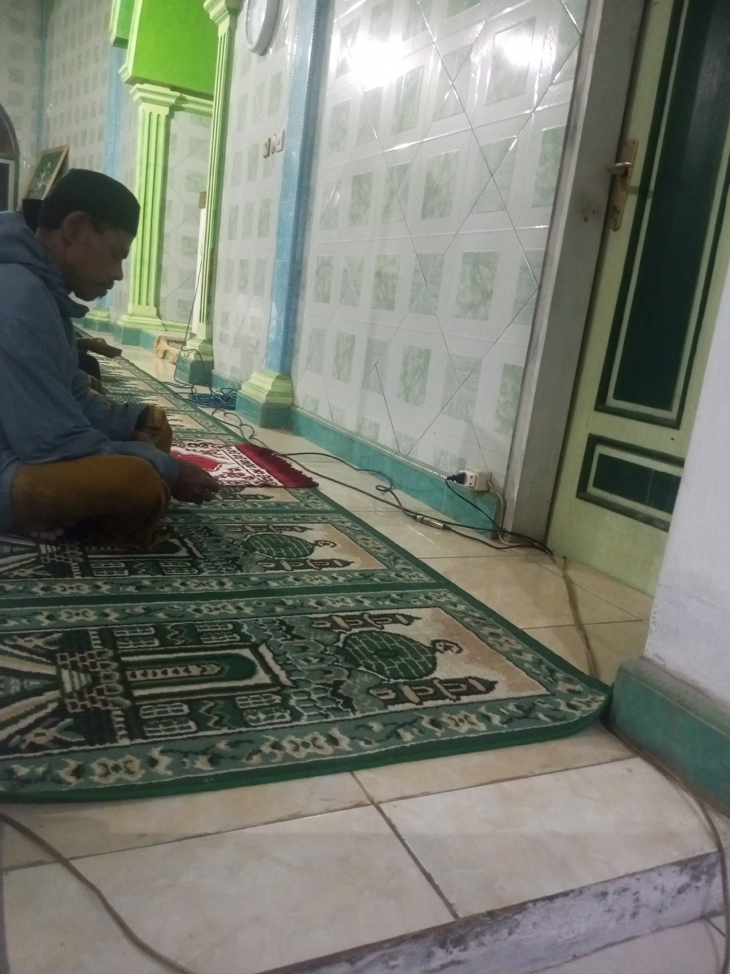 A man, wearing a blue jacket and brown pants, is sitting on a prayer rug in a mosque. He is facing away from the camera and appears to be praying. The prayer rug has a green and white pattern. The walls of the mosque are tiled with a light green and white pattern. There are green pillars in the background. A door with a dark green frame is visible on the right side of the image. The floor is tiled with light brown tiles. Electrical cords and an outlet are visible near the wall. This scene is likely within the city of Andongsili, Indonesia.